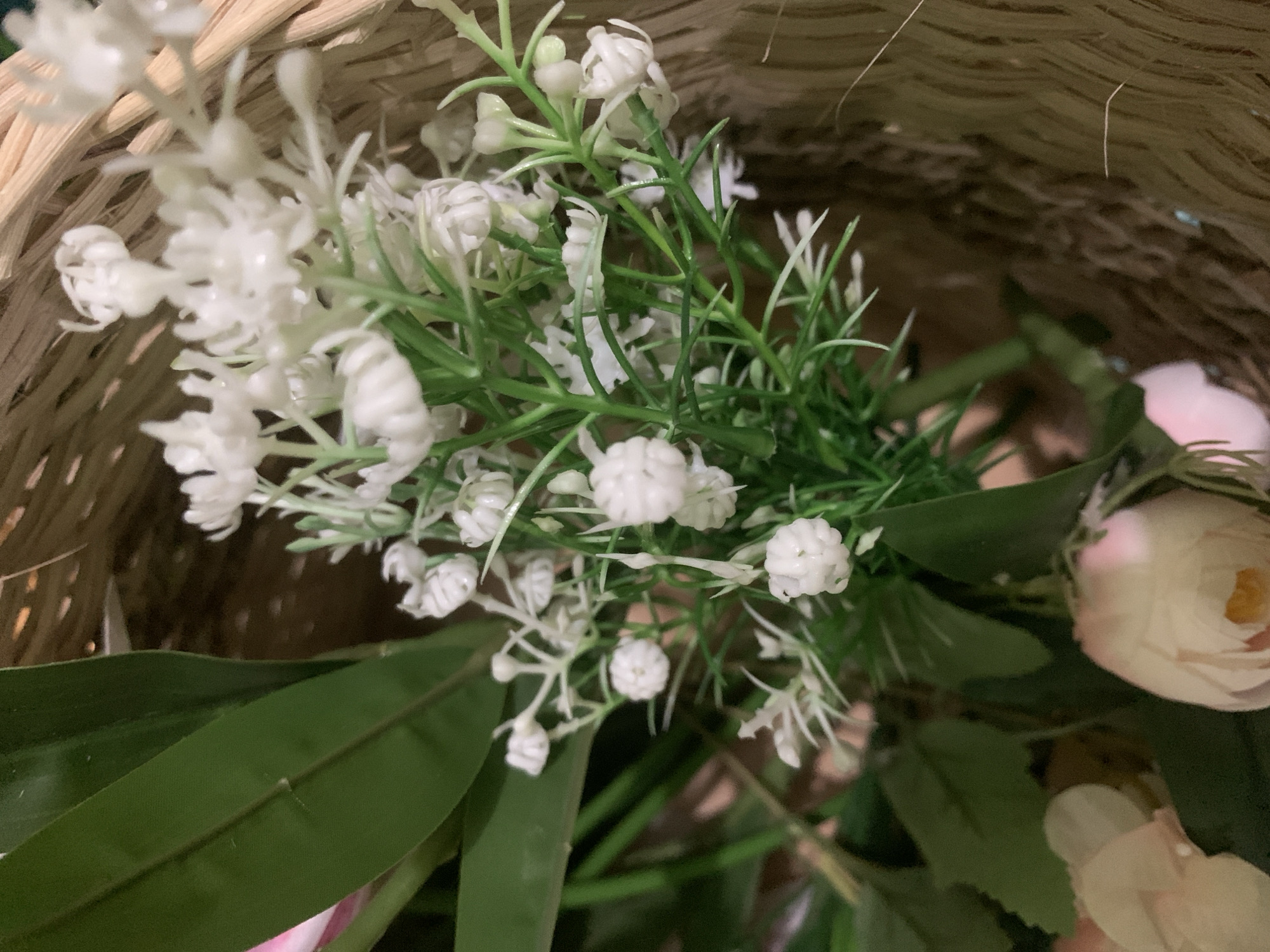 The image is a close-up shot of artificial flowers, primarily white, with green foliage, arranged in a woven basket. The flowers are delicate and clustered, resembling baby's breath or small blossoms. Some larger, pale pink roses are partially visible in the lower left and bottom center of the frame. The background consists of the textured weave of the basket and a dark, out-of-focus area. The lighting appears to be soft, highlighting the texture of the flowers and basket. There is no discernible indication of the city of Kuala Pembuang, Indonesia within the image.