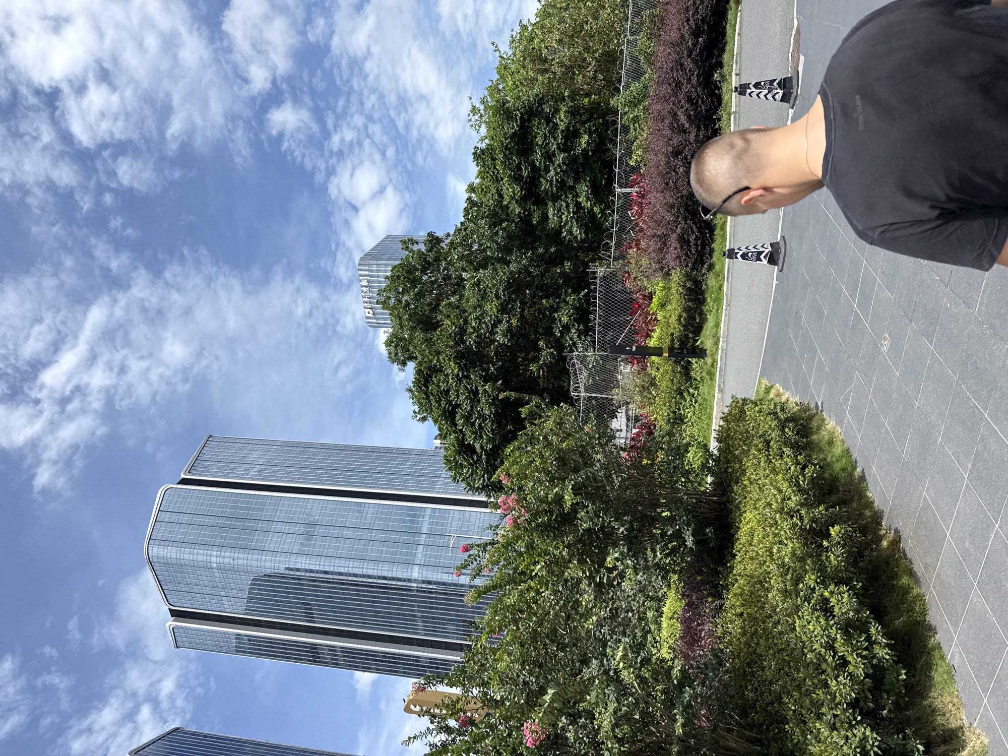 In Huangpu District, China, on a sunny day with scattered clouds, the image captures a modern cityscape dominated by tall, glass-fronted skyscrapers. In the foreground, a person wearing a black t-shirt and sunglasses perched on their head is walking away from the camera, their back to the viewer. The ground is paved with grey tiles arranged in a grid pattern. To the left, lush green bushes and trees with pink flowers form a vibrant border, contrasting with the urban architecture behind. Further back, a chain-link fence encloses a strip of landscaping featuring red and purple foliage. Two black and white traffic cones are visible on the pavement, suggesting ongoing work or restricted access in the area. The overall scene depicts a blend of natural elements and contemporary urban development.