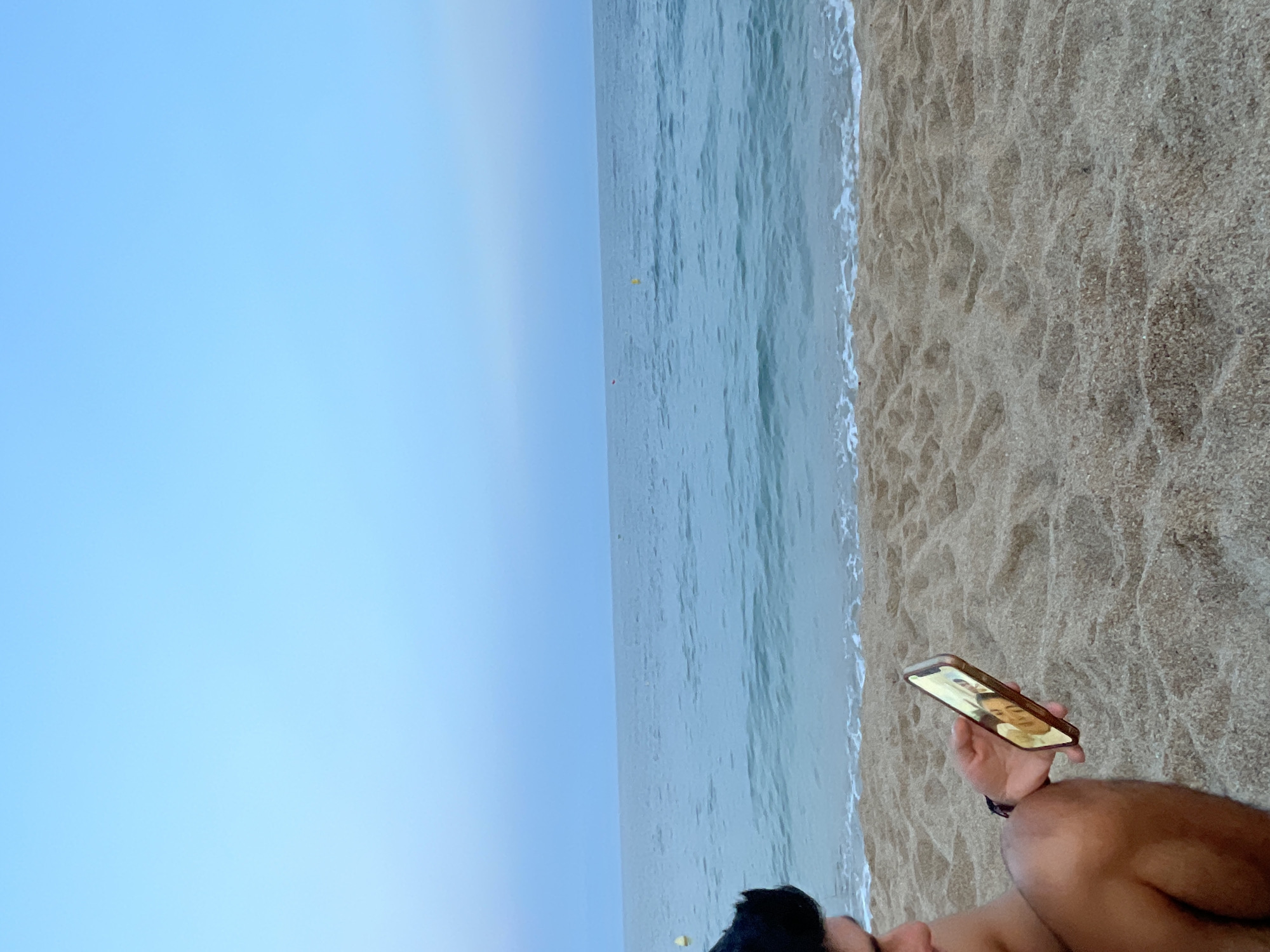 A young person sits on a sandy beach in Canet de Mar, Spain, looking at a smartphone. The sky is a clear, pale blue, transitioning to a slightly deeper blue at the horizon. The sea is a tranquil turquoise, with gentle waves lapping at the shore. The sand is a warm, light brown, showing the texture of small grains and subtle undulations. The person, partially visible from the side, has dark hair and is wearing dark shorts. They are holding a smartphone in their right hand, the screen displaying a picture of two individuals. The scene suggests a peaceful moment of leisure by the Mediterranean Sea.