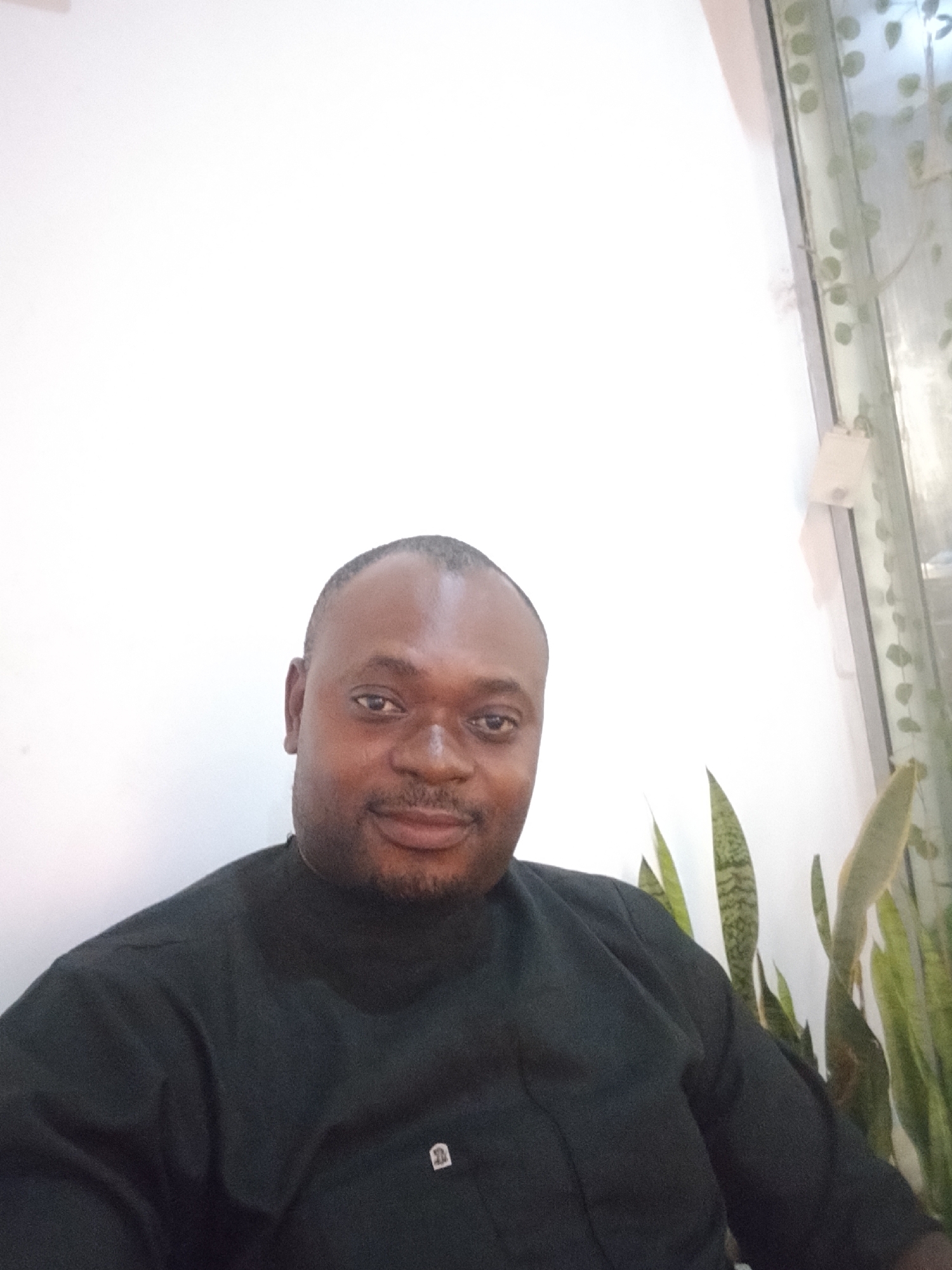 A man sits indoors in Gwarinpa, Nigeria. He is wearing a dark-colored, long-sleeved shirt. Behind him is a white wall and a window with sheer curtains. Green, leafy plants are located near the window.