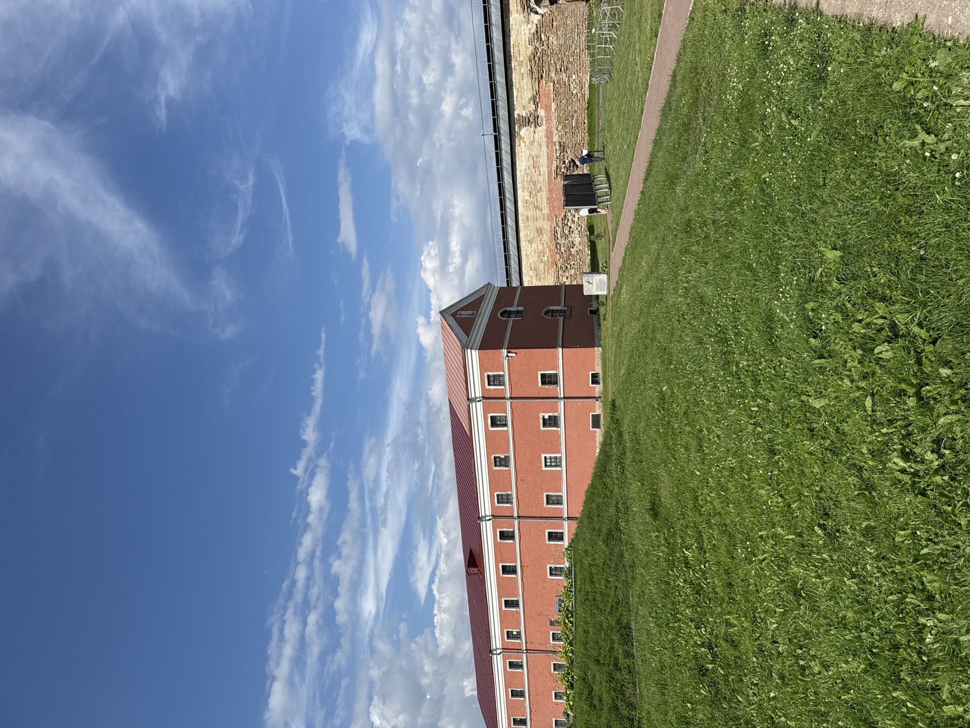 In Shlisselburg, Russia, an exterior view captures a red brick building with a red roof and numerous windows, situated behind a grassy hill. Adjacent to this building is a weathered stone wall topped with a metal railing. The sky above is a vibrant blue, dotted with scattered white clouds. In the middle ground, a paved path leads through a grassy area where two individuals are observed standing near informational displays. The scene is bathed in daylight, suggesting fair weather.