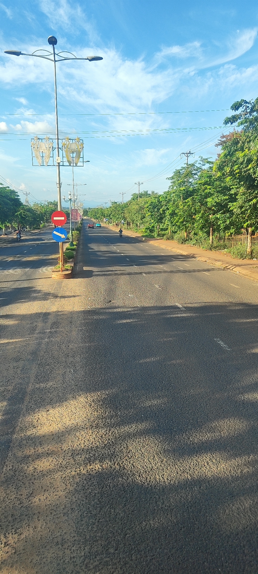 This is a photograph of a wide, paved road in Nhơn Hòa, Vietnam, lined with mature green trees on both sides. The sky above is blue with scattered white clouds. Streetlights are visible along the road, and power lines stretch overhead. In the foreground, shadows from the trees are cast across the asphalt.
Several vehicles are present: a red car and a light blue car are driving in the distance on the right side of the road, and a motorcycle is visible on the left side. A person is riding a motorcycle on the right side of the road. Two traffic signs are mounted on a pole near the left side of the road: a circular red sign with a horizontal white bar, indicating "No Entry," and a blue circular sign with a white arrow pointing left, indicating "Mandatory Turn Left." The road appears to be a main thoroughfare with clear lane markings. The overall scene suggests a relatively sunny day in a semi-urban or rural area.