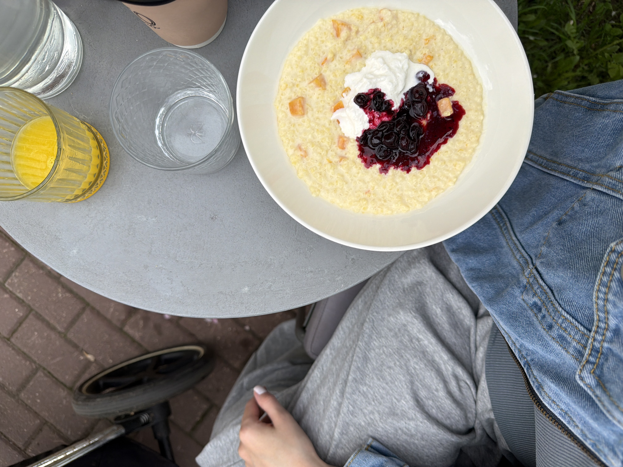 The image shows a top-down view of a scene on a light gray round table. A white bowl filled with porridge topped with what appears to be cream and dark berry compote sits on the table. Next to the bowl are a glass of orange juice, a clear glass of water, a disposable cup (partially visible), and another glass of water.
The bottom of the image shows a person seated at the table, wearing gray sweatpants and a blue denim jacket. A small portion of a black stroller wheel is visible in the lower left.
The location is outdoors, evidenced by brick paving visible in the lower left and greenery in the upper right corner. The exact location in Khimki, Russia, cannot be determined from the image alone.