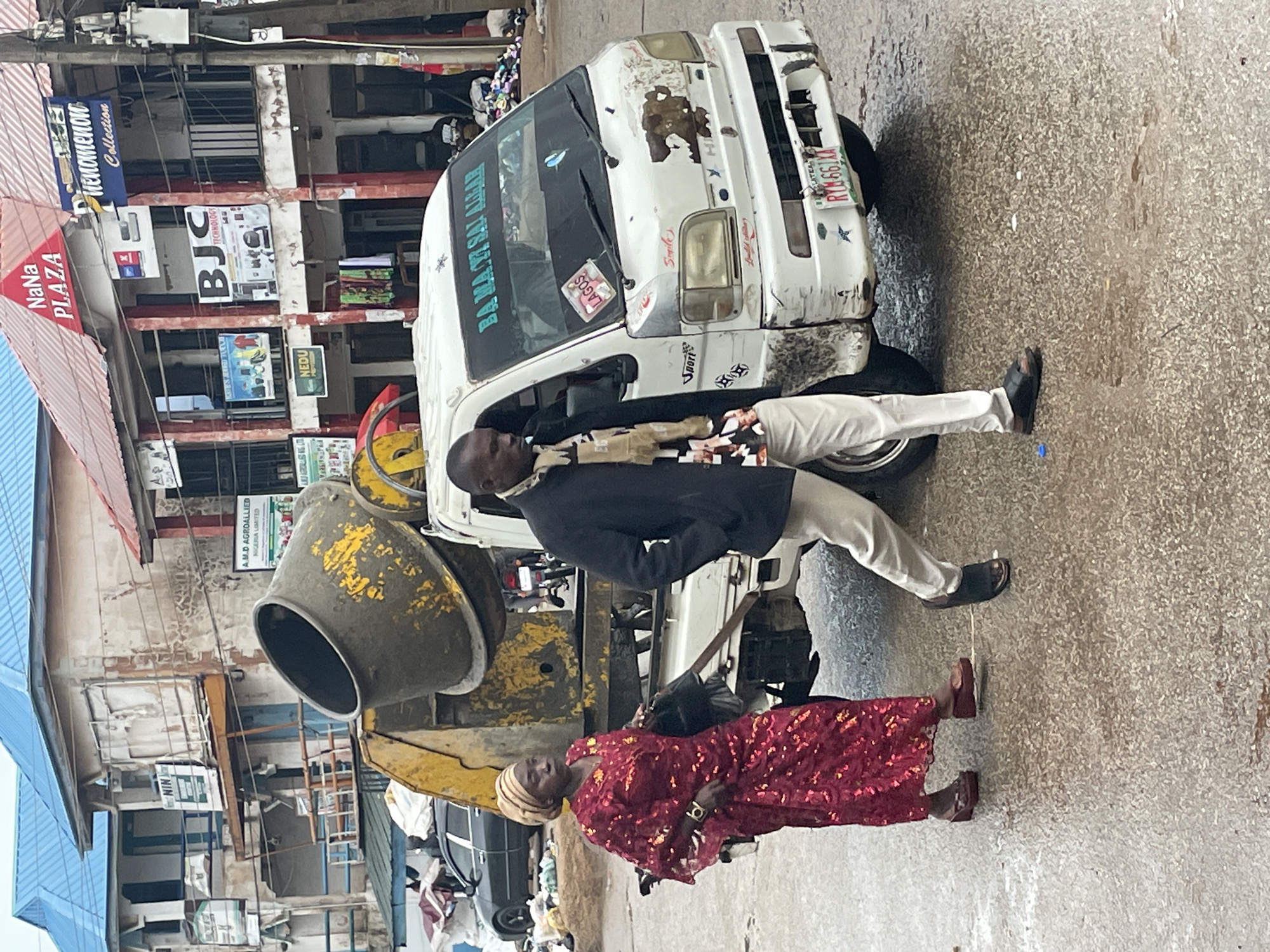 In Bukuru, Nigeria, a scene unfolds on a wet, paved street in front of commercial buildings. A large, yellow cement mixer is mounted on the back of a white utility truck. Two individuals walk past the truck. A woman on the left wears a vibrant, shimmering red patterned dress and a beige headscarf, accessorized with a dark bag and dark sandals. Beside her, a man on the right is dressed in a black jacket, light-colored pants, and black sandals. He carries a patterned scarf. The truck itself is adorned with various stickers, including one that reads "LAGOS," and its windshield displays the phrase "BA MA YI SAL ALHAM." The buildings behind the truck feature signs for "NaNa PLAZA" and "Phenomenoro Collection." The environment appears to be a busy urban area with a mix of commercial activity and a general sense of unkemptness, evident by debris visible in the background.
