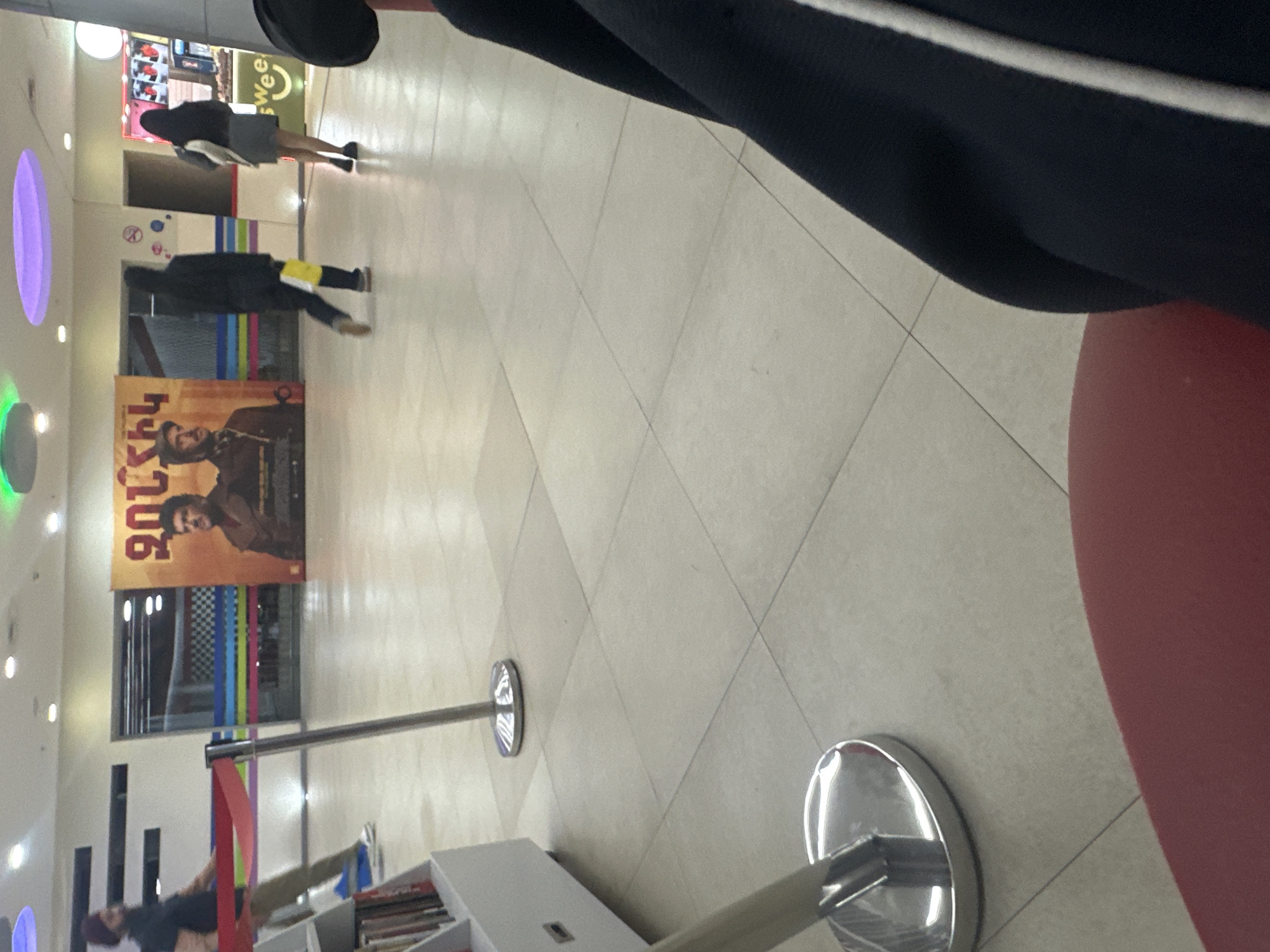 The image captures an indoor public space, likely a mall or cinema lobby, viewed from a low perspective as if the photographer is seated. The frame is dominated by a light-colored tiled floor with square patterns, leading towards the background. In the immediate foreground, part of a person's leg or lap in dark clothing with a white stripe is visible, alongside a reddish surface, suggesting a seat.
Mid-ground shows several individuals. Two people walk away from the camera; one wears dark clothing with a yellow item on their back, and the other, further ahead, has a dark top and light-colored skirt, carrying a white object. Towards the far left, another person in dark attire and light pants stands near white shelving units, possibly displaying books or merchandise.
The background features light-colored walls adorned with various elements. A large, prominent poster on the left displays two men, one with a beard wearing a brown jacket. Partial text "UCKY" is visible, along with text in a South Asian script above it. To the right of the poster, a window or glass partition features colorful vertical stripes in blue, green, yellow, pink, and red. A green sign with the partial word "Swee" and a stylized 'e' resembling a smiling face can be seen further right. The ceiling is dotted with recessed circular lights, some emitting purple or green glows.
Several silver stanchions with red retractable belts are placed throughout the area, indicating crowd control or queuing. The overall scene suggests a bright, modern, and orderly public environment.