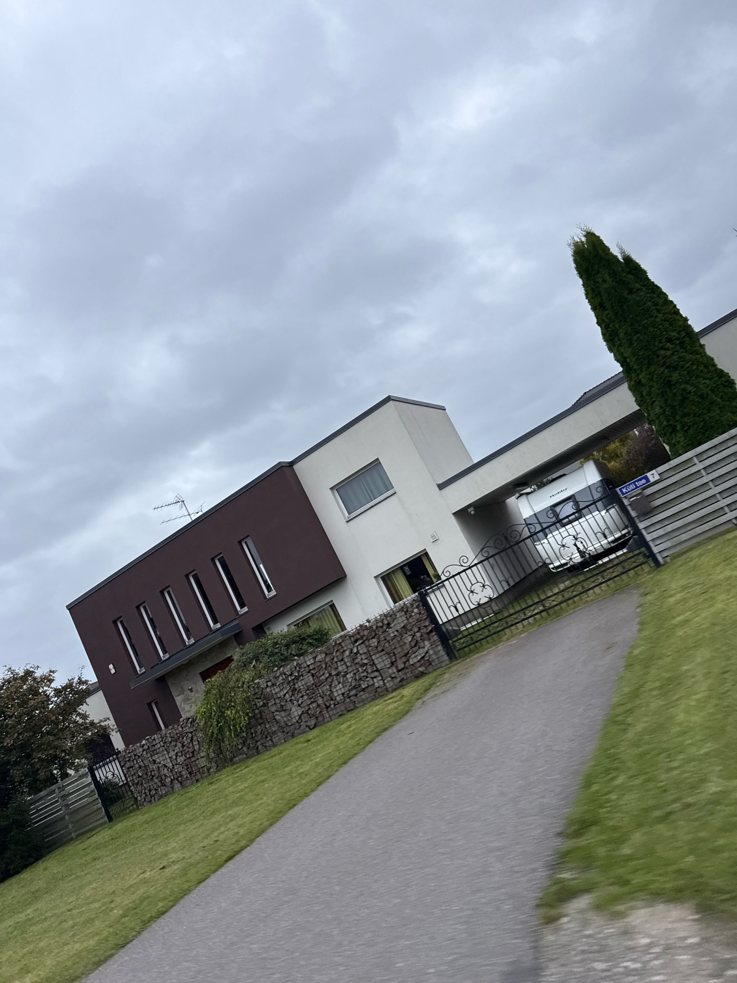 The image depicts a modern house in Peetri alevik, Estonia, on a cloudy day. The house has a distinctive architectural style with contrasting dark brown and white sections, featuring a series of narrow, vertical windows on the dark section. A stone wall borders the property. A paved driveway leads to a carport where a white recreational vehicle (RV) is parked. A black metal fence with decorative elements stands in front of the RV and the house entrance. To the right of the house, a tall, slender evergreen tree stands prominently. A street sign with the text "Külvi tee 7" is visible near the fence. The surrounding area includes a well-maintained green lawn and other fencing. The scene is captured from a slightly tilted perspective, giving a dynamic feel to the composition.