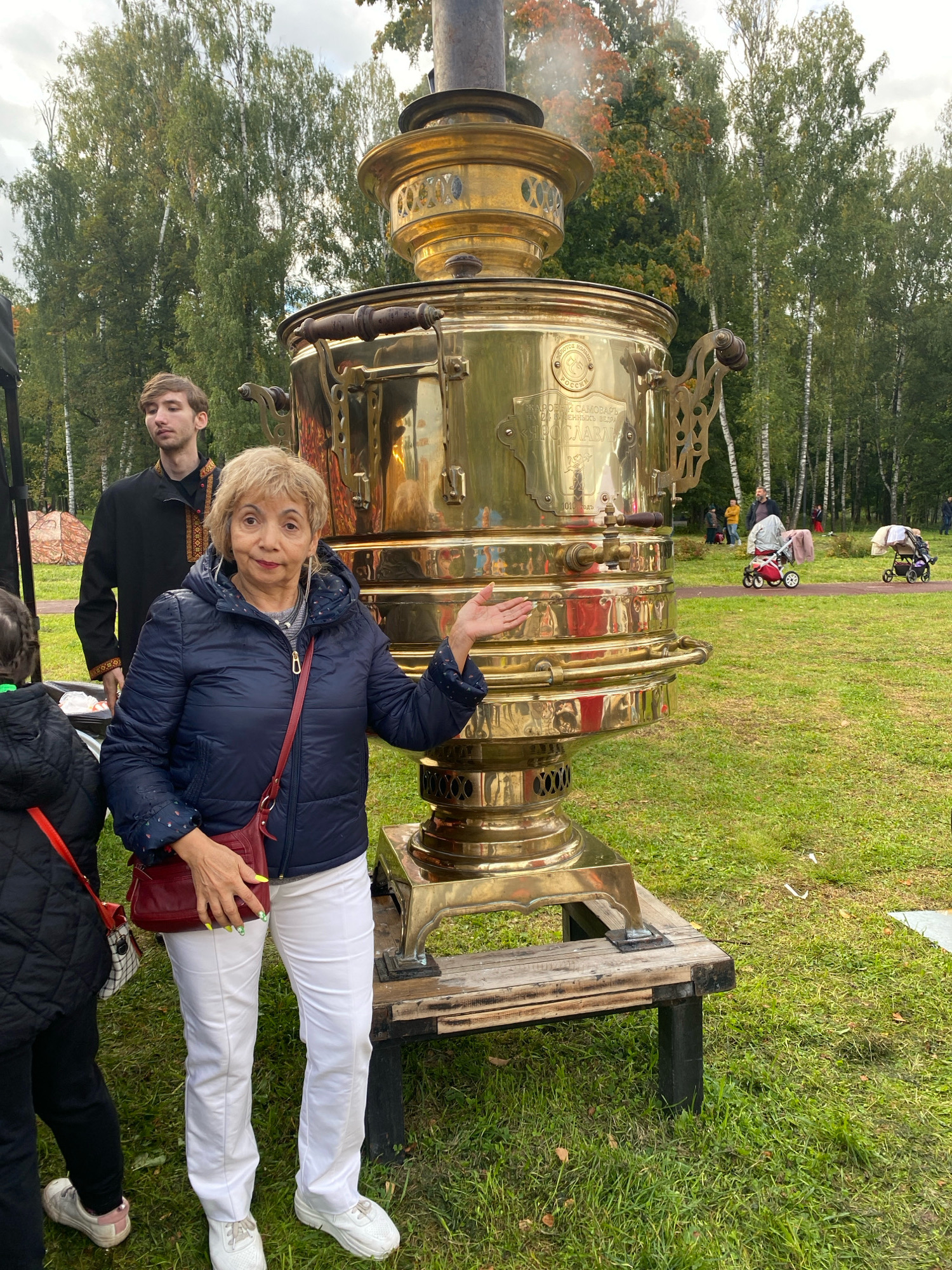 An elderly woman with blonde hair and a blue jacket stands in front of a large, ornate brass samovar in an outdoor setting with lush green grass and trees. She is holding a red purse and gesturing with her left hand. Behind her, a young man in traditional attire stands to the left, and further in the background, other people and strollers are visible on a path. The samovar is steaming, indicating it is in use. The scene appears to be a park or open-air gathering space.