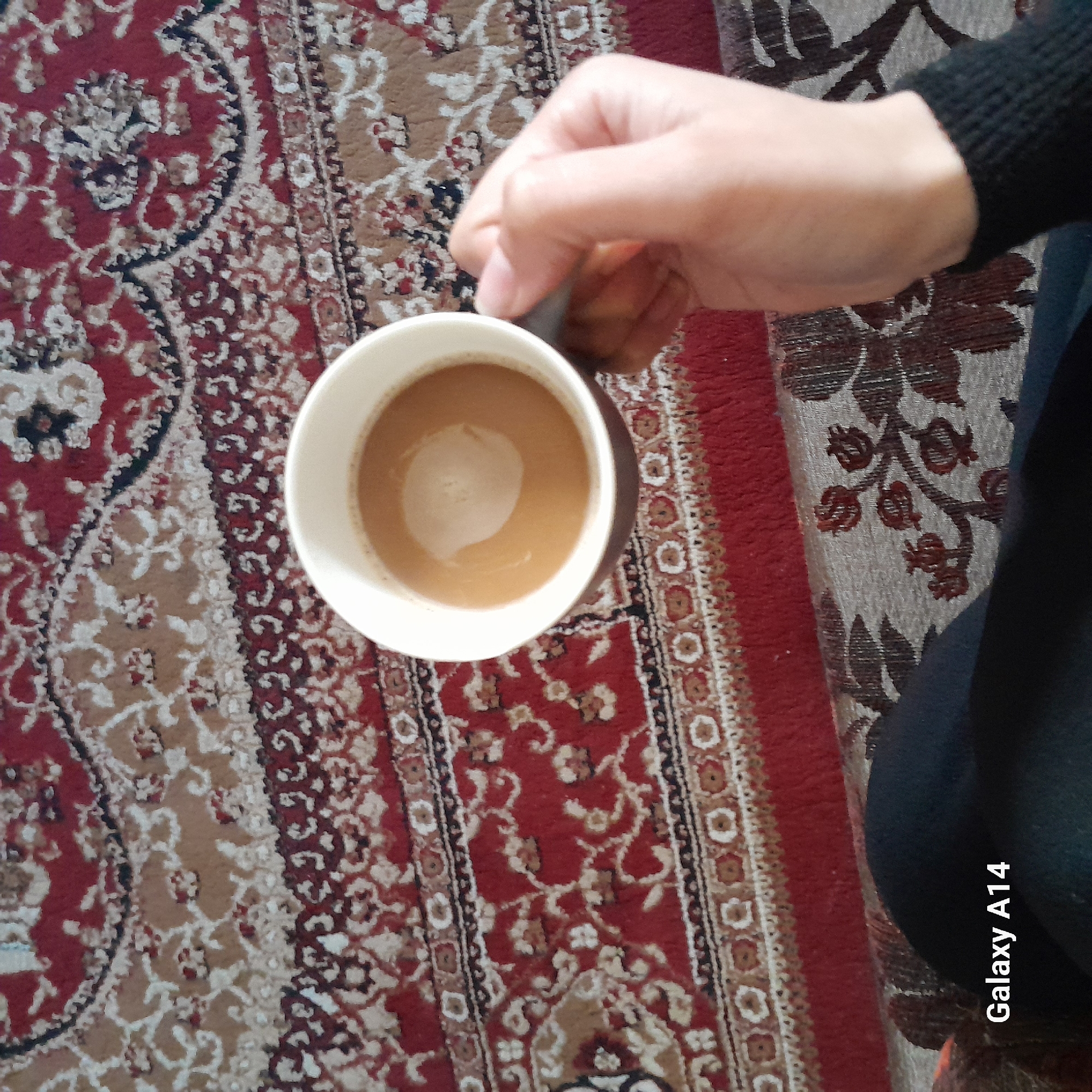 A person holds a mug filled with a light-brown liquid, possibly coffee or tea with milk, against the backdrop of an ornate rug. The rug features repeating patterns in shades of red, tan, and black. The person's hand and part of their arm, clad in a black long-sleeved garment, are visible. The bottom right corner of the image shows a blurred black object and the text "Galaxy A14".
