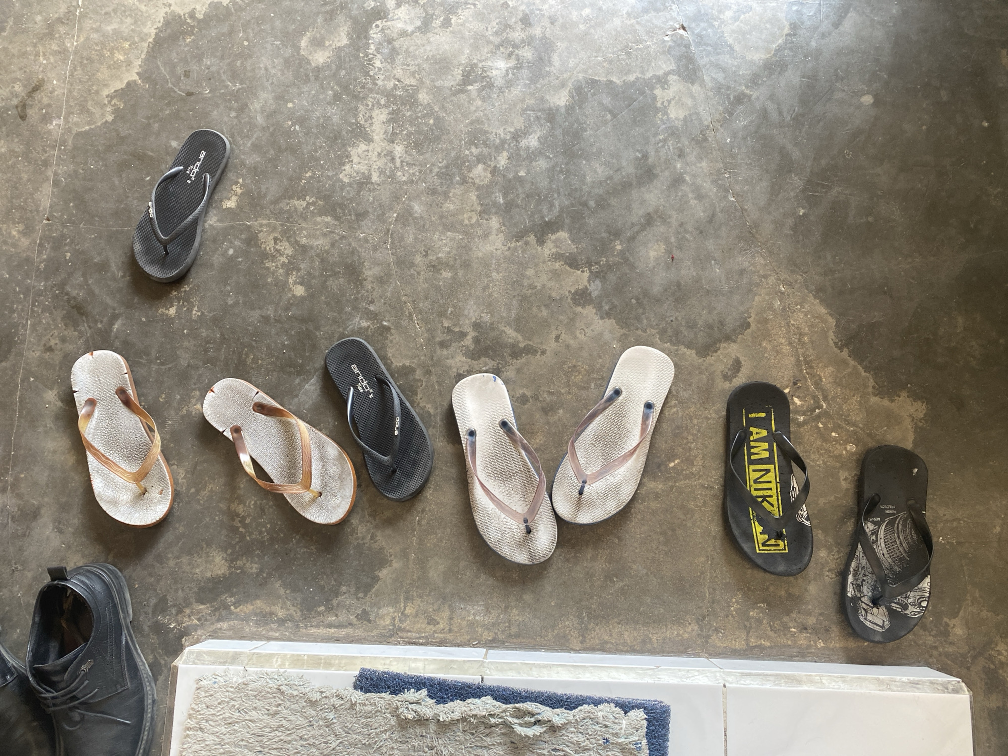 A pair of black dress shoes rests on a textured mat next to a concrete floor. Scattered across the floor are several pairs of flip-flops. One pair is light brown with darker brown straps. Another pair is white with translucent straps. Two pairs of black flip-flops are also present; one has the text "I AM NIKON" in yellow on the sole, and the other features an image of a camera lens. The scene appears to be an outdoor or semi-outdoor area, possibly a porch or entryway, in Kotakulon, Indonesia.