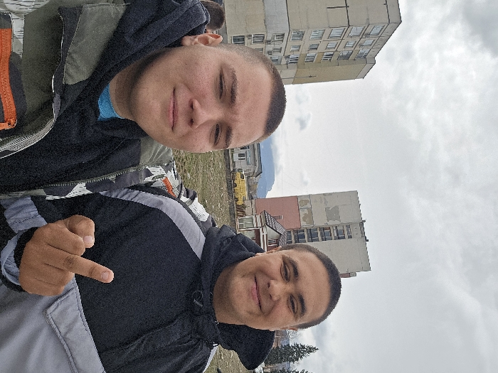 Two young males are depicted in an outdoor urban setting in Samokov, Bulgaria, under an overcast sky. The male on the left, with light skin and a buzz cut, wears a blue shirt, a dark hooded sweatshirt, and an olive green/camouflage pattern jacket, looking at the camera with a slight smile. The male on the right, with a darker skin tone and a buzz cut, wears a black and gray two-tone hooded jacket, smiling broadly at the camera. His right hand is raised, with the middle finger extended. In the background, two tall, weathered, multi-story concrete residential buildings are prominent; the rightmost building features a faded pink lower facade. A small yellow van is visible in the mid-ground. Distant mountains and coniferous trees are present on the horizon and to the right, respectively. The immediate ground appears to be an unpaved, uneven area.