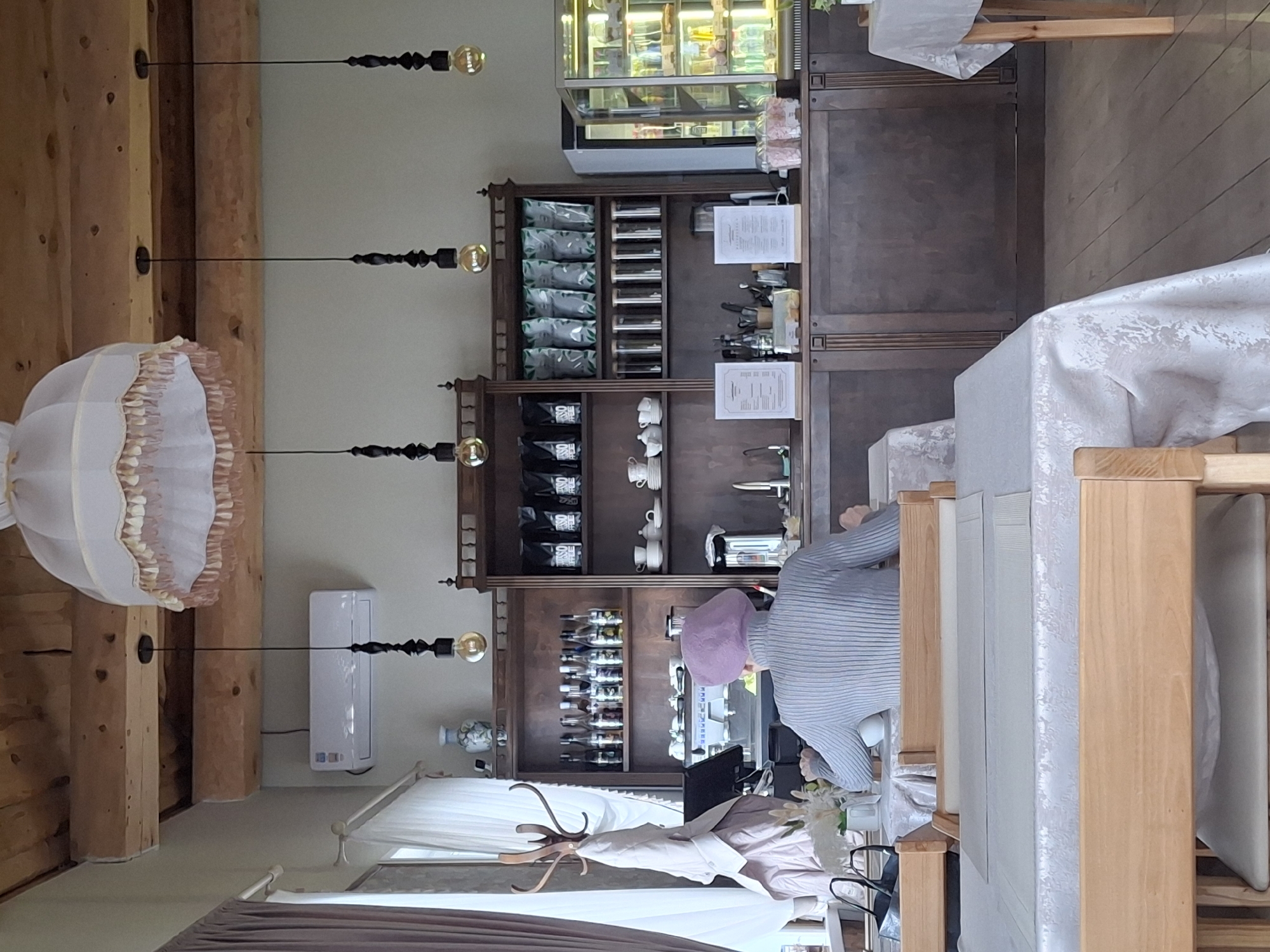 This image depicts the interior of what appears to be a cafe or restaurant in Sarapul, Russia. The scene is dominated by a long, dark wooden counter or shelving unit that stretches across the back and right side of the room. On the top section of this unit, a glass-fronted display refrigerator contains various items, possibly pastries or desserts. Below, shelves are stocked with numerous packages, some featuring the text "OSHO," likely coffee bags. White cups and saucers are neatly stacked, and a collection of bottles (possibly syrups or spirits) is also visible. An espresso machine and other beverage preparation equipment are set up on the counter, suggesting a barista station.
One person is seen behind the counter, facing away from the camera, wearing a light blue ribbed top and a purple head covering. They appear to be engaged in preparing a drink or working at the coffee machine.
In the foreground and to the right, several wooden tables are covered with white tablecloths, some with light grey placemats, and paired with wooden chairs with light grey seats, set for potential diners.
The left side of the room features rustic wooden beams running horizontally, from which several minimalist pendant lights with exposed bulbs hang. A large, ornate, light-colored fabric chandelier or lampshade is also suspended in this area. A white air conditioning unit is mounted on the wall. Further into the left foreground, a wooden coat rack holds a jacket, and a white sheer curtain partially covers a window. The overall ambiance suggests a blend of rustic and refined decor. The time of day is not discernible.