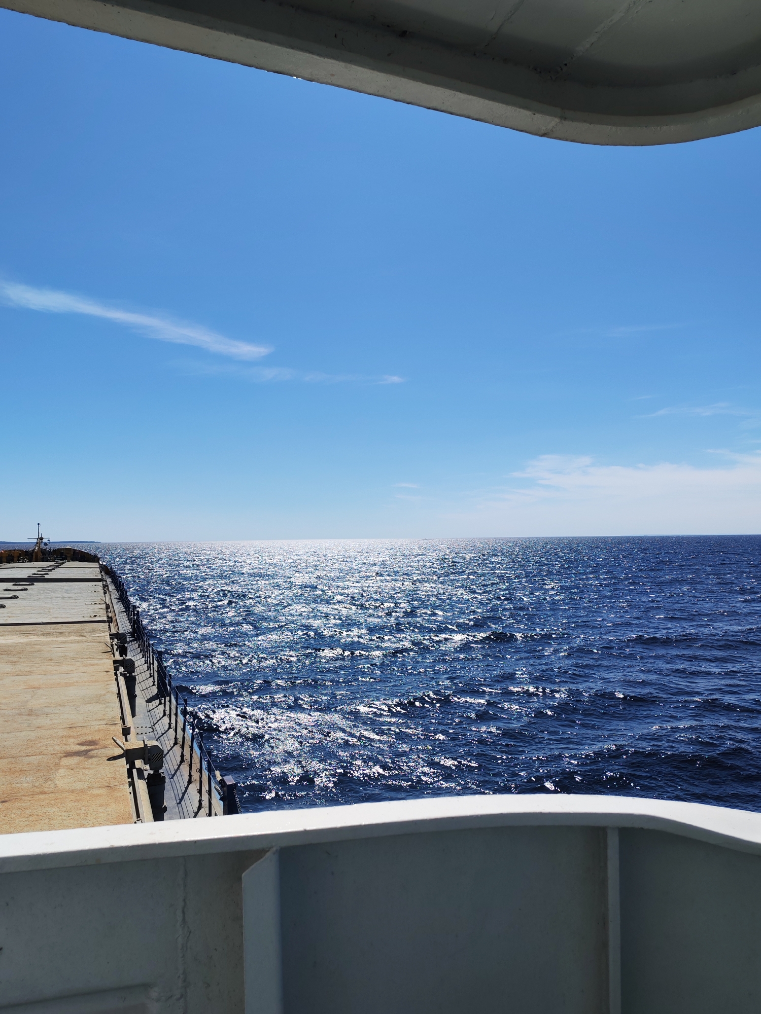 The image captures a view of the sea from what appears to be a pier or a ship's deck. The sea is a deep blue, with sunlight reflecting off the rippling surface, creating a bright, shimmering effect. A long, grey structure, possibly a pier, extends into the water on the left side of the frame. It is made of concrete and metal railings line the edge closest to the water. The sky is a clear, bright blue with a few wispy white clouds visible. The foreground shows a portion of a white structure, also likely part of a ship or pier, which frames the view of the sea and sky. There are no people visible in the image. This scene could be located near Shal'skii, Russia, a location known for its coastal access and maritime activities.