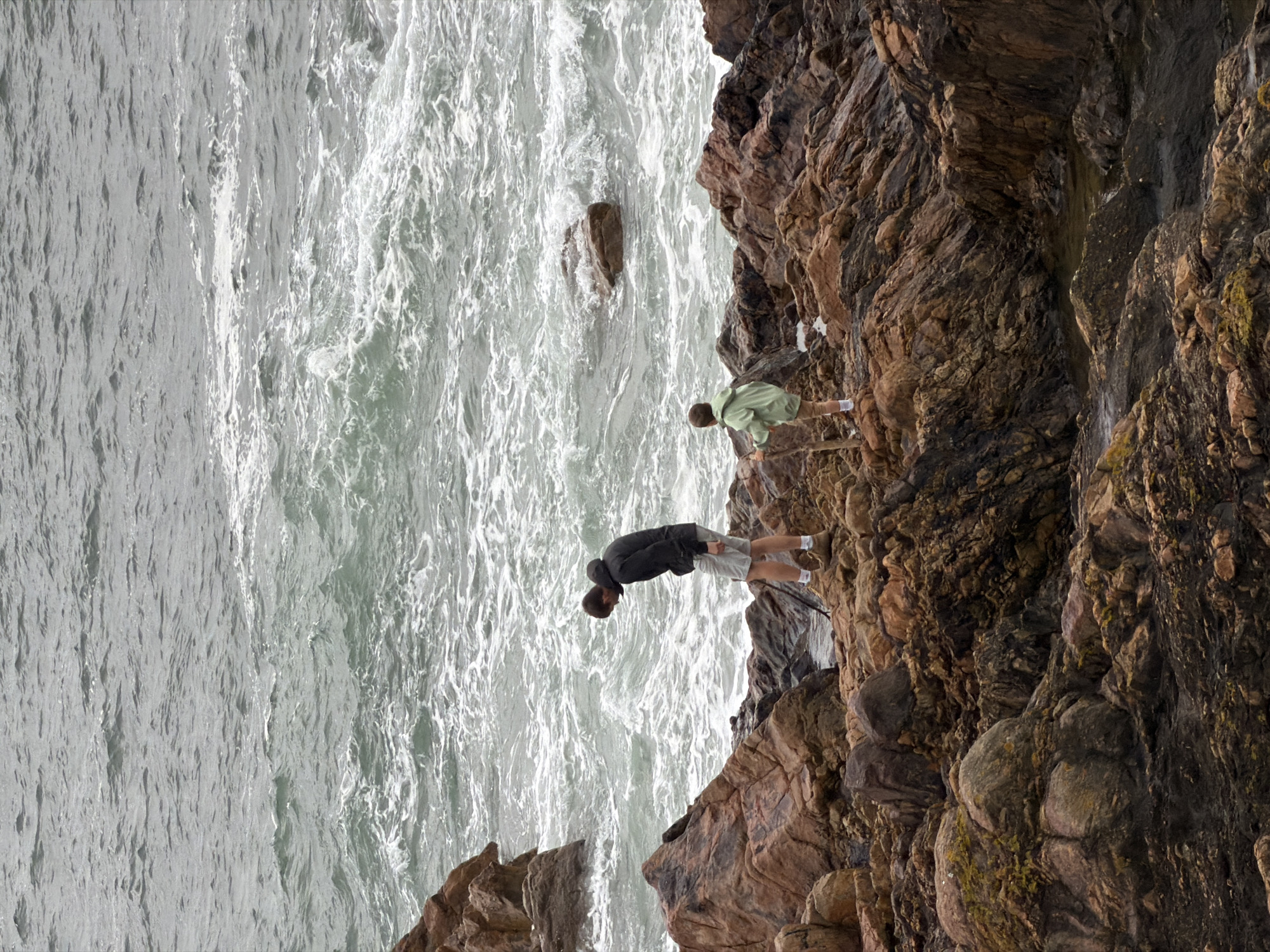 Two individuals are seen on a rocky coastal area, likely in Porto, Portugal, as suggested by the landscape's characteristics. One person, wearing a dark jacket and light-colored shorts, appears to be leaning over the edge of the rocks towards the sea. Another individual, wearing a light green shirt and khaki shorts, is standing further back. The rocky terrain is rugged, consisting of various shades of brown and grey. The sea is turbulent, with white foamy waves contrasting against the darker grey water. A large rock stands alone within the body of water.