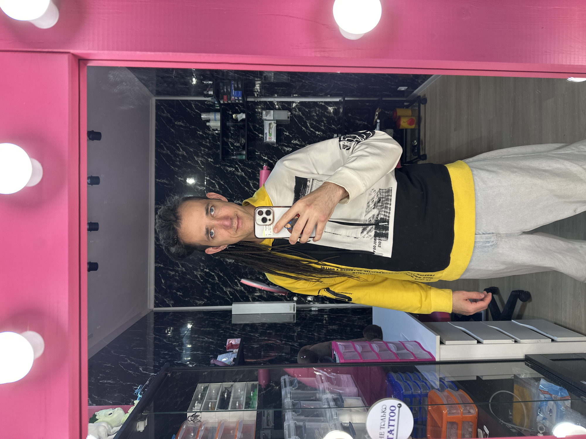 A man with shoulder-length dark braided hair is reflected in a mirror with a pink frame and light bulbs. He is holding a smartphone and taking a selfie. He is wearing a sweatshirt that is white, black, and yellow, and light-colored jeans. The background behind the mirror shows a room with black marble-patterned walls. Shelves are visible in the background, stocked with various items. Below the mirror, there's a glass display case containing small containers and packaging, including a white circular sticker that reads "НЕ ТОЛЬКО TATTOO". Based on the Russian text and the general appearance, the location is likely a tattoo parlor in Saratov, Russia.