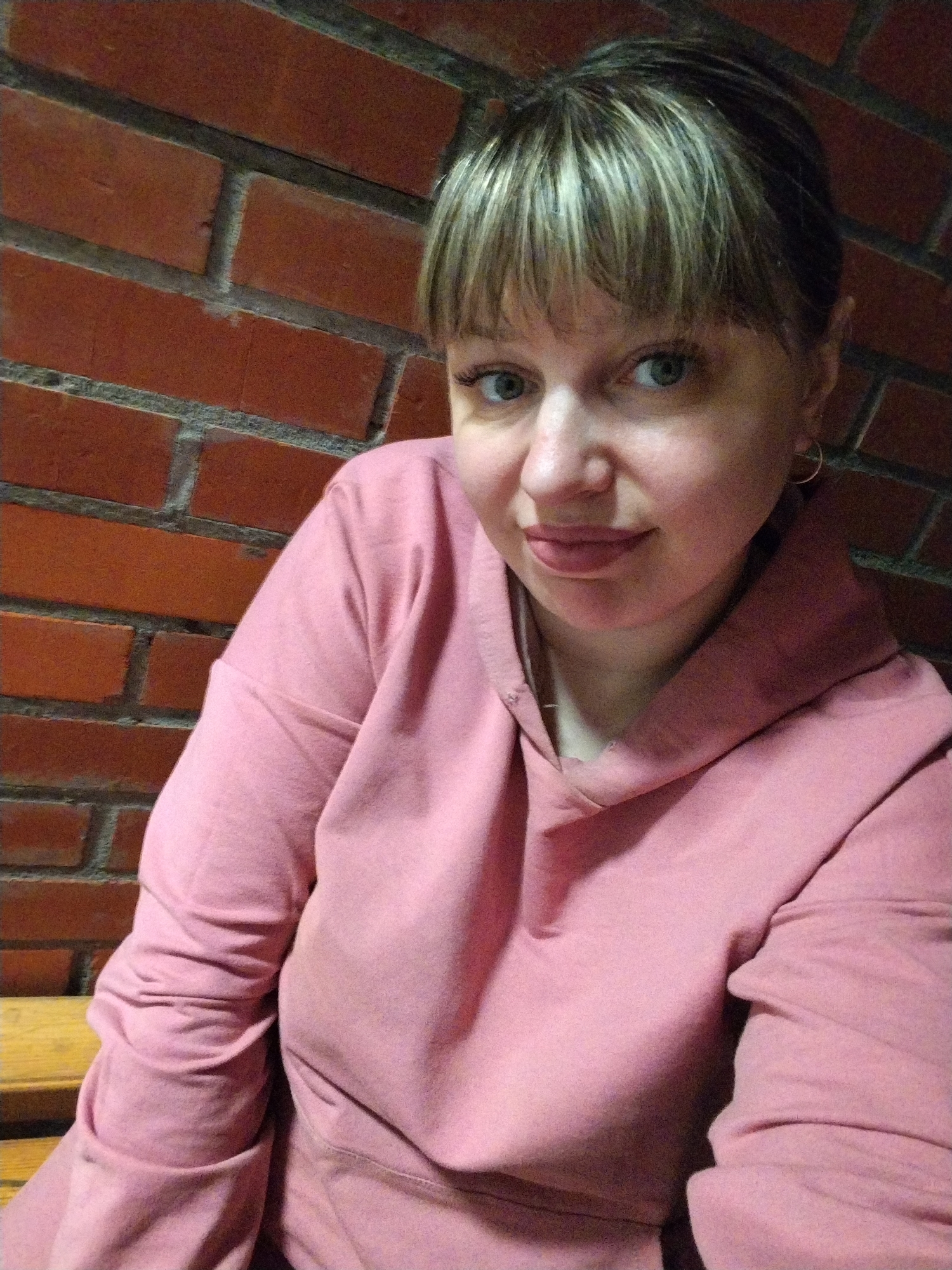 A woman is featured in a medium close-up portrait, looking directly at the camera with a neutral to slightly contemplative expression. She has light skin, striking green-blue eyes, and light brown hair with blonde highlights, styled with bangs across her forehead. A small, golden hoop earring is visible in her right ear. She is wearing a soft, pink hooded sweatshirt (hoodie), which covers her from the chest up.
The background consists of a reddish-brown brick wall with light gray mortar lines, suggesting an indoor or sheltered outdoor setting. In the lower left corner of the frame, a segment of a light-colored wooden surface, possibly a bench or a table, is visible. The lighting in the image appears somewhat dim, characteristic of an indoor environment or evening hours. There is no specific activity taking place other than the woman posing for the photograph. No text is visible in the image. The provided location context is Шебунино, Russia.