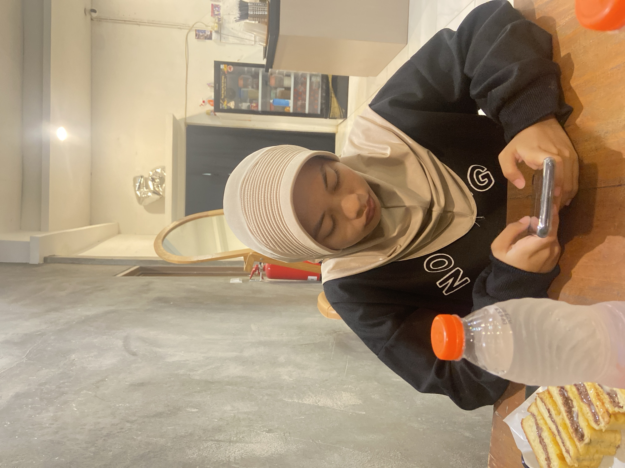 The image shows a young person wearing a light brown hijab and a black sweatshirt with white lettering, sitting at a wooden table. They are looking down at a smartphone held in their hands. To the left of the person is a bottle of water with an orange cap, and further left is a plate of toasted bread with a filling. The background shows a concrete wall, a mirror, and a doorway leading into a room with shelves stocked with various items. The scene appears to be an indoor establishment in Sekar Mawar, Indonesia.