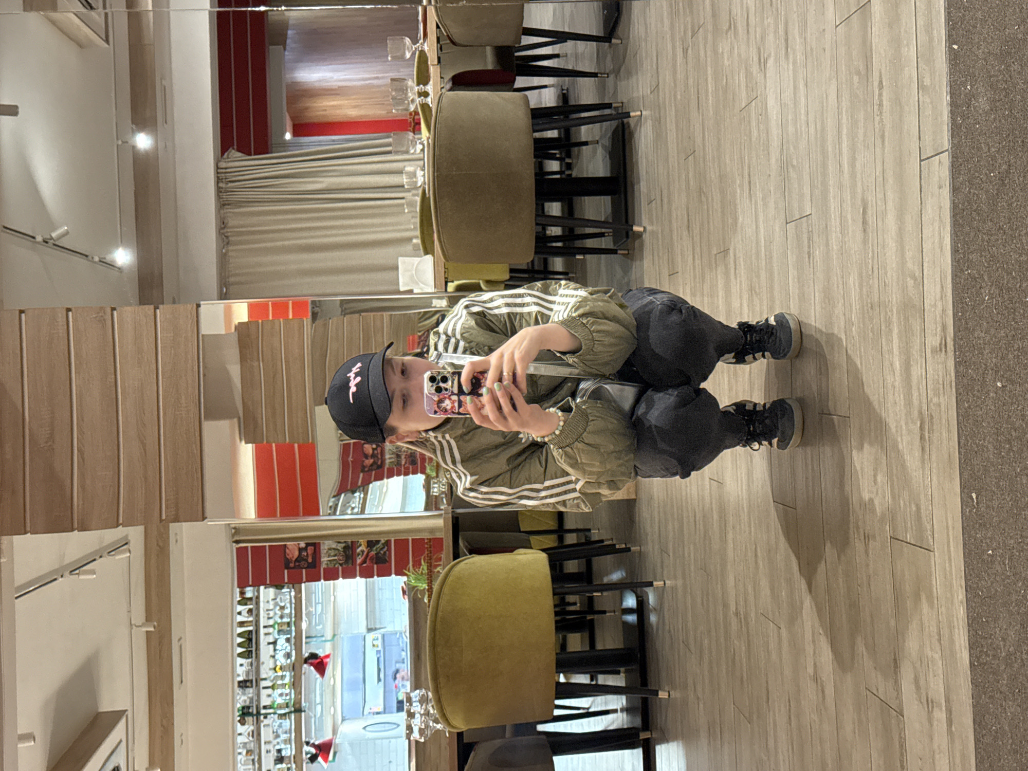 A mirrored image shows a person in a restaurant or café. The person is crouching and holding a smartphone, presumably taking a picture of themself in the mirror. The individual is wearing a black baseball cap, a green jacket with white stripes, and dark pants. The shoes are dark-colored sneakers. The reflection in the mirror reveals a well-lit interior with chairs, tables, and a bar area stocked with bottles. The decor suggests a modern or contemporary style. Based on the presence of Russian text on the phone case and other visual clues, the location could be in the city of Moscow, Russia, but this is not definitively verifiable from the image.