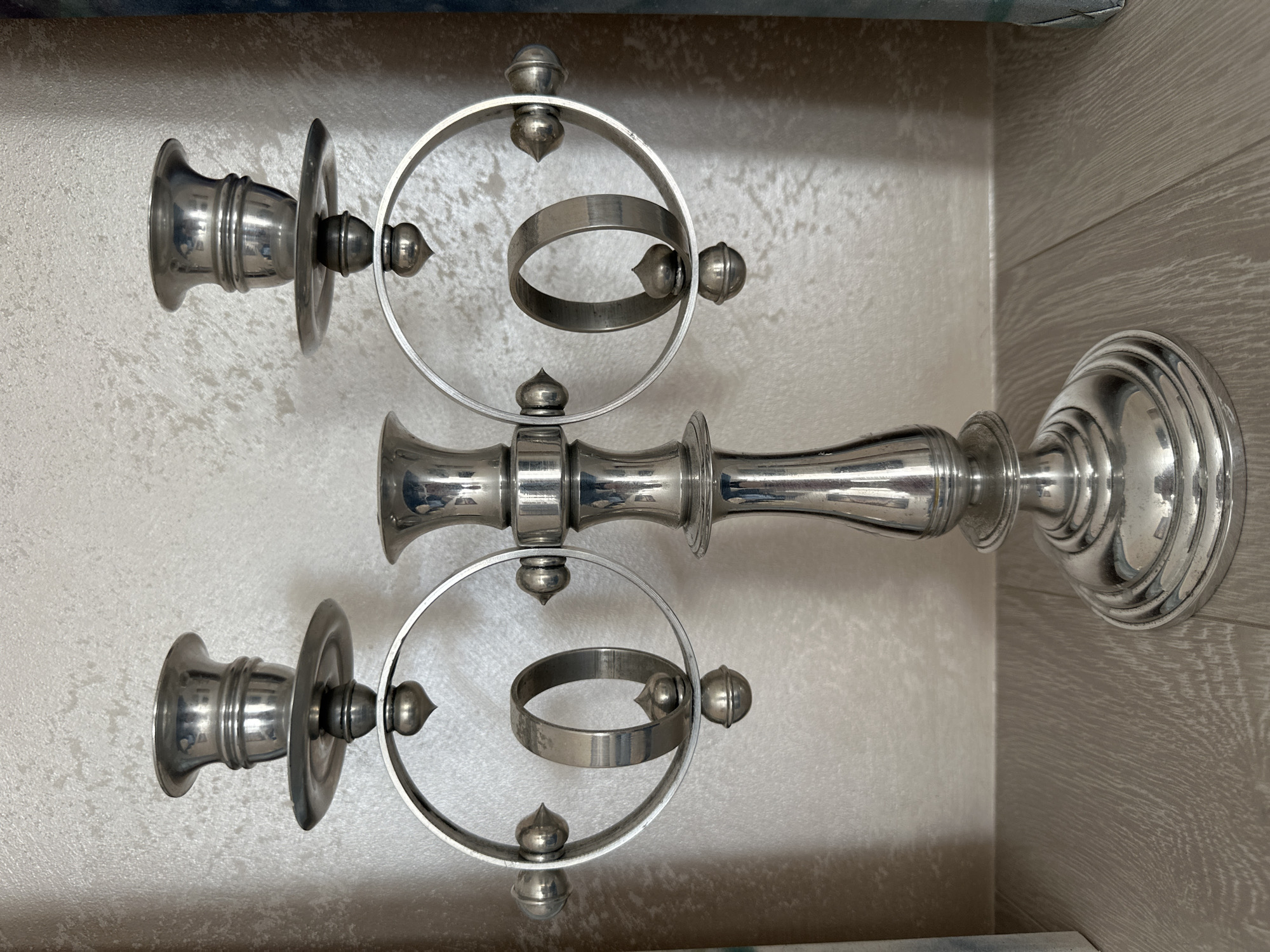 A polished silver candelabra with two candle holders sits on a light-colored wooden surface. The candelabra has a central stem with decorative bands, leading to two arms, each supporting a candle holder. Attached to these arms are circular frames, each containing a smaller, suspended ring. The background is a textured, light-colored wall. There are no people or animals present in the image. The scene appears to be indoors, likely in a domestic setting, with no discernible connection to the city of Minsk, Belarus.