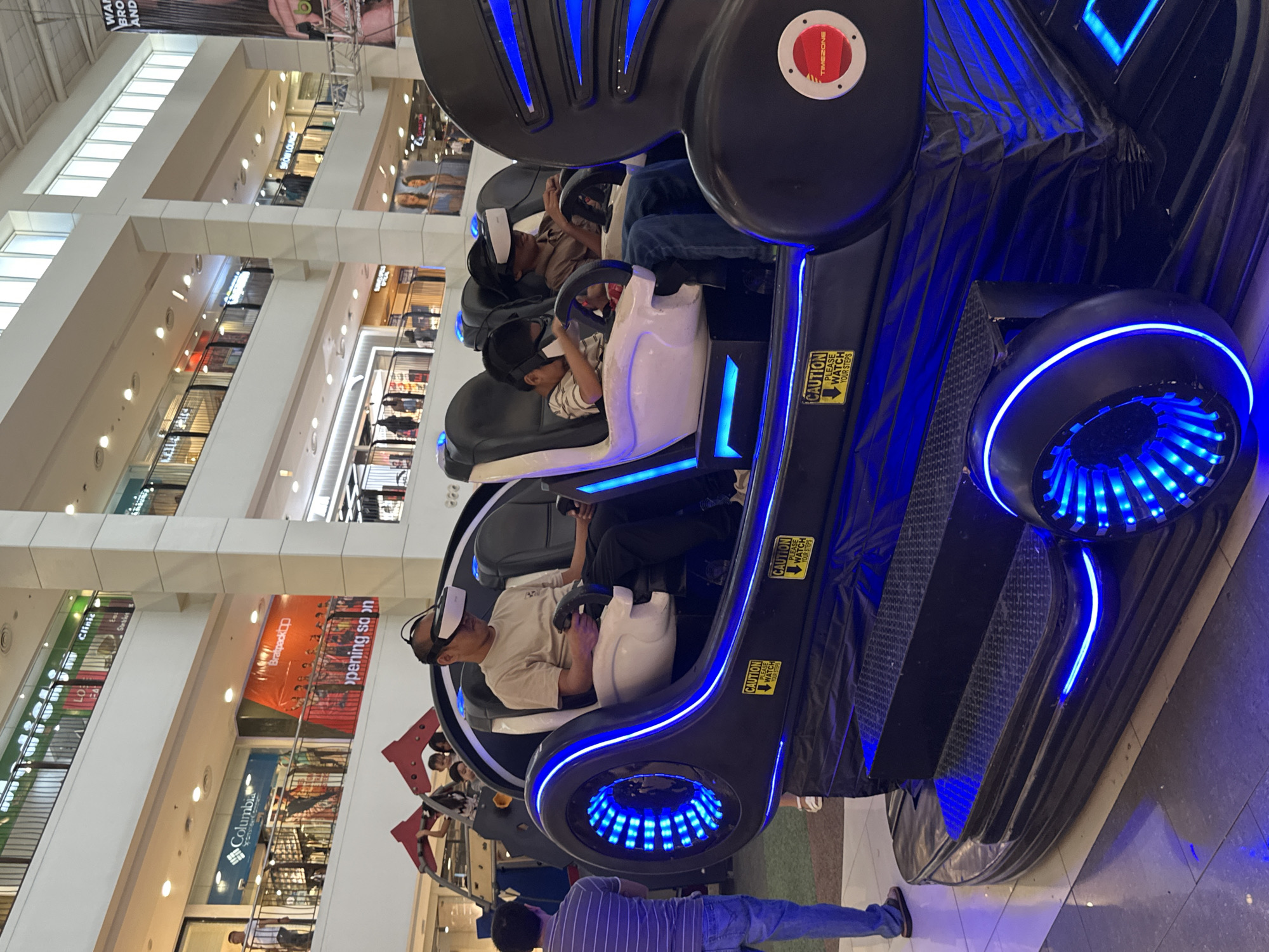 The image depicts an indoor scene, likely a shopping mall in Davao City, Philippines, given the context. In the foreground, a black and blue illuminated motion simulator ride with multiple seats is prominent. Three individuals are seated within the ride, all wearing virtual reality headsets and holding controllers, indicating they are engaged in a VR experience. One adult male is in the front left seat, and two young males occupy the seats behind him. The ride features blue LED lighting accents along its exterior and a "TIMEZONE" logo on its side. A man in a striped shirt and jeans stands to the left of the ride, facing away from the camera. In the background, the multi-level interior of a mall is visible, with shops and escalators. Stores like "Columbia" and a banner reading "Bratherp BO opening soon" are discernible, suggesting a commercial setting. The overall atmosphere is casual, with people participating in entertainment activities.