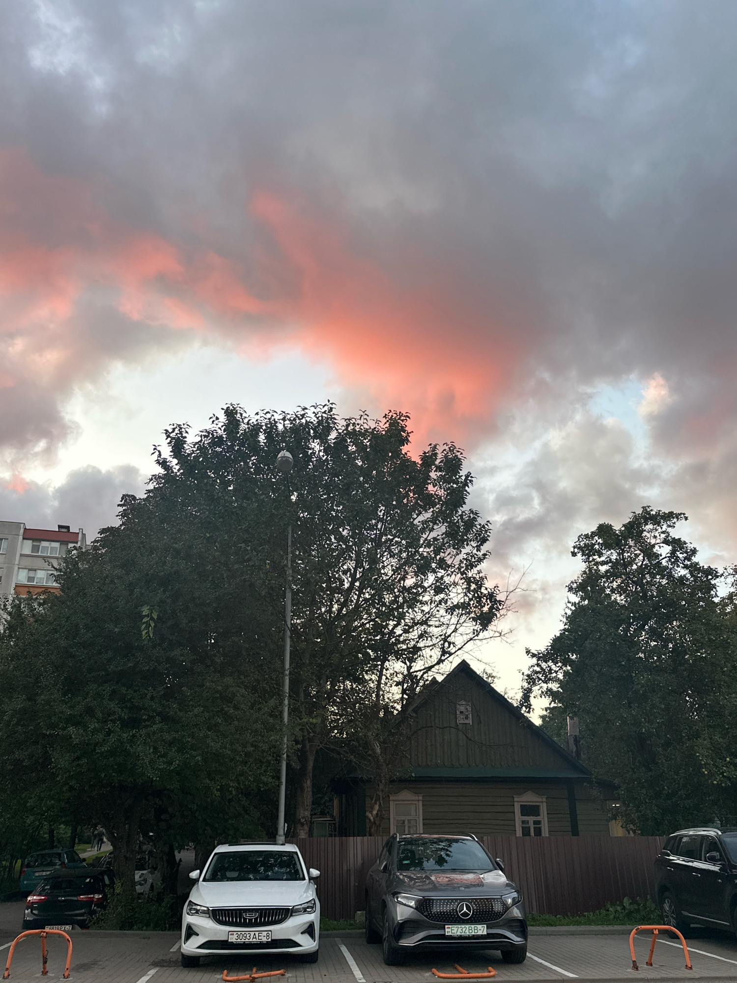 The image depicts a parking lot in Minsk, Belarus, with a striking sunset in the background. In the foreground, two cars are parked: a white Geely with license plate "3093 AE-8" and a dark gray Geely with license plate "FE732BB-7". Behind the cars, a traditional wooden house with a dark green facade stands next to a dense cluster of trees. To the left, a multi-story apartment building is visible. The sky is filled with dramatic clouds, some of which are illuminated by the setting sun, casting a warm, pinkish glow. A street lamp is also present in the scene, positioned near the trees. The ground is paved with interlocking bricks, characteristic of urban parking areas. The overall scene suggests an evening in a residential area of the city.