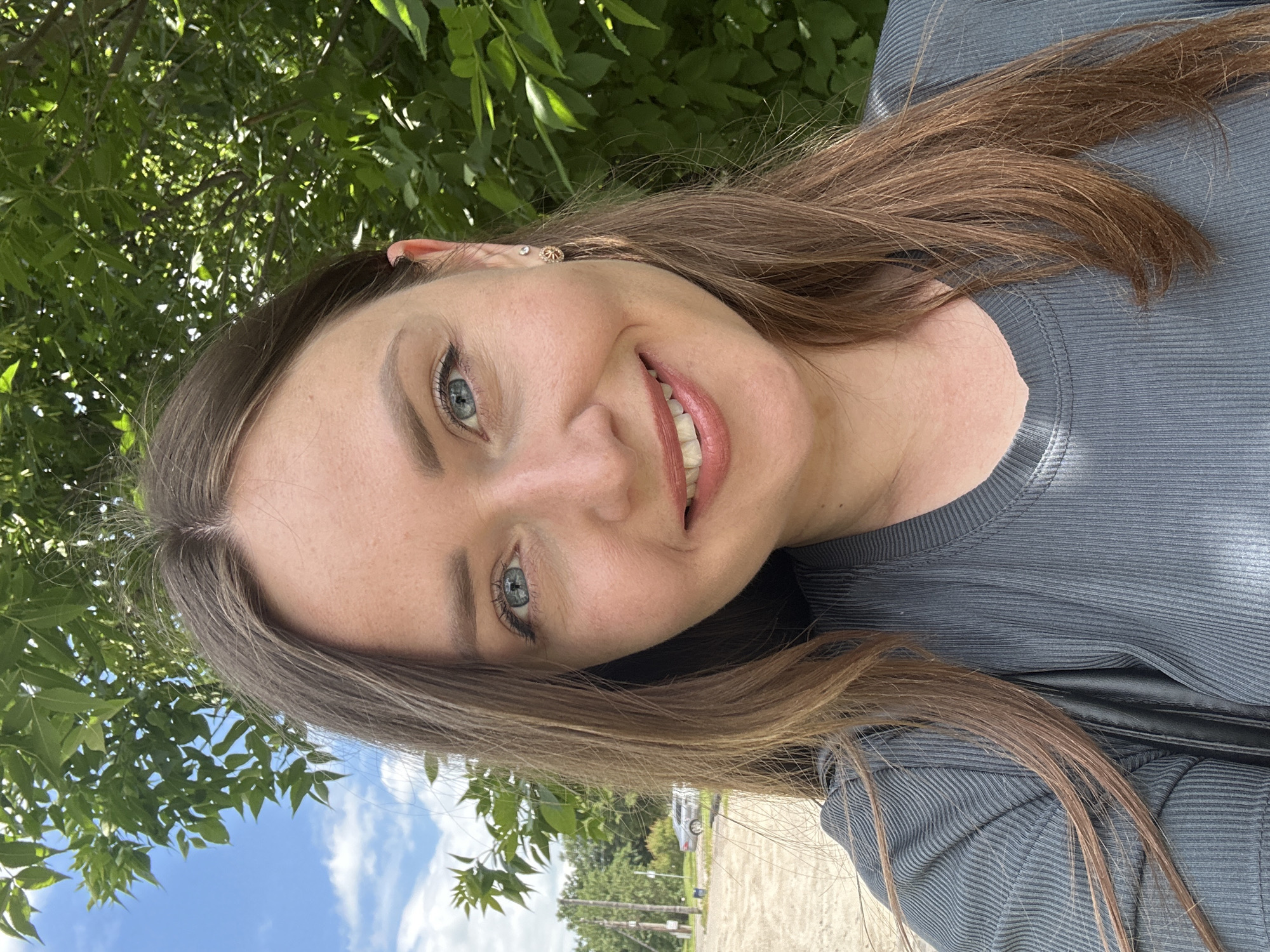 This is a slightly tilted, close-up selfie of a woman taken outdoors. She has long, brown hair that falls across her shoulder and chest. Her face is turned towards the camera, and she is smiling. She has blue eyes and fair skin. She is wearing a grey, ribbed, long-sleeved shirt.
The background consists of lush green leaves from a tree overhead, with a bright blue sky with white clouds visible in the distance. A glimpse of a paved area and some distant trees and a parked car suggest an outdoor, possibly rural or suburban, setting. The lighting indicates it is daytime, likely a sunny day given the brightness. There is no visible text in the image.
The image appears to be a casual, personal photograph, possibly taken during a sunny day outdoors, in Bila Tserkva, Ukraine.