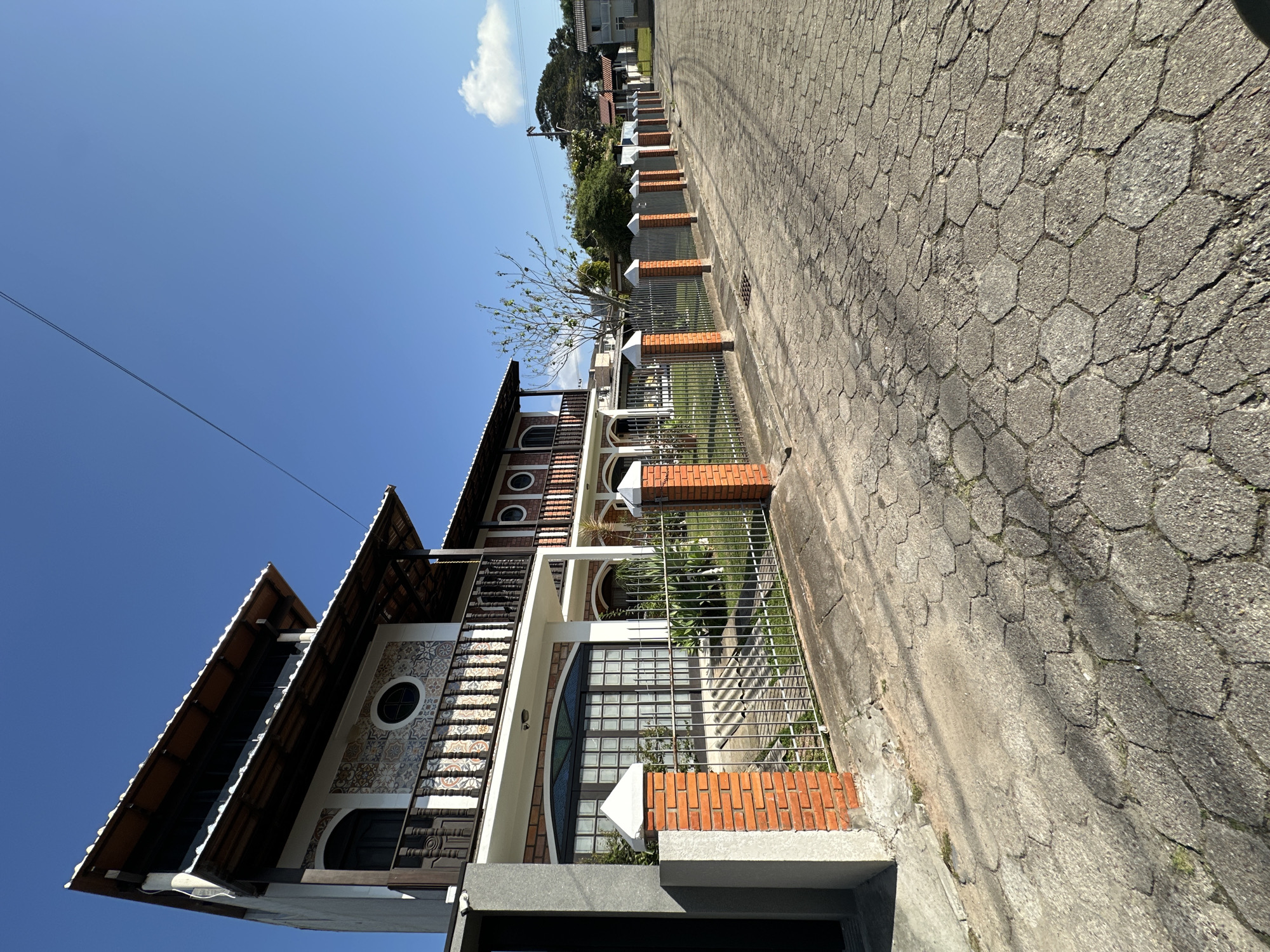The image depicts a street scene in the city of Florianópolis, Brazil, under a clear blue sky. The foreground is dominated by a hexagonal-patterned cobblestone road, showing signs of wear and tear. A metal fence with brick pillars lines the left side of the road, separating it from a property. Behind the fence, a two-story building with a distinct architectural style is visible. The building features a pitched roof with wooden beams, multiple balconies with ornate railings, and decorative tile work on its facade. The ground floor has large glass doors. Further down the street, other houses and trees are visible, suggesting a residential area. The overall impression is one of a sunny day in a semi-urban environment. There are no people or vehicles present in the image.