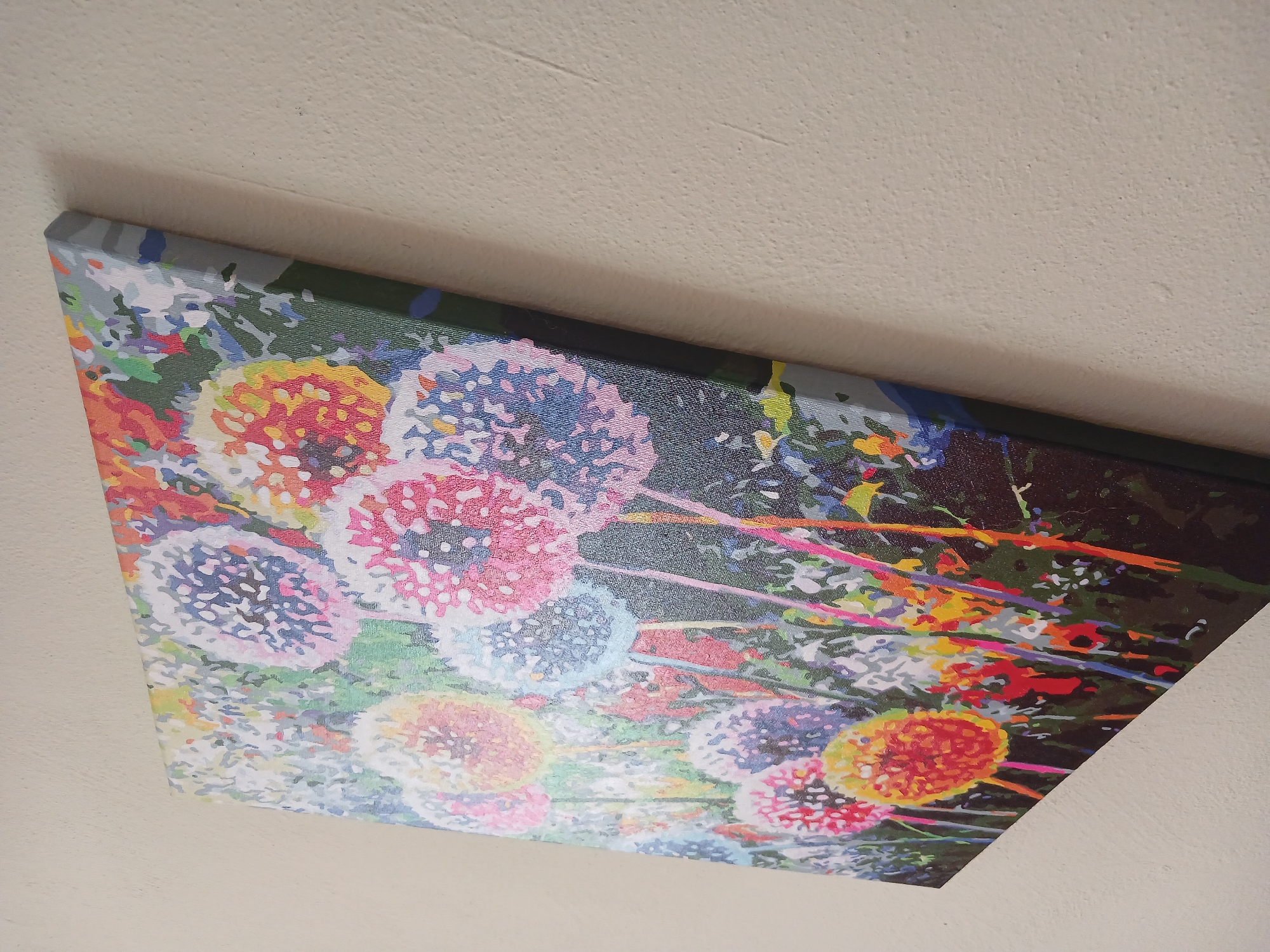 The image shows a colorful painting of dandelions, possibly a paint-by-numbers artwork, mounted on a wall. The painting is predominantly in shades of pink, blue, yellow, and orange, depicting stylized dandelion seed heads against a dark green background. The edges of the canvas are visible, and it appears to be stretched over a frame. The painting is leaning against a textured, light-colored wall, casting a subtle shadow. The composition suggests the artwork is part of an indoor setting, though no other objects or architectural features are clearly visible. The image was taken in Mamaivtsi, Ukraine.