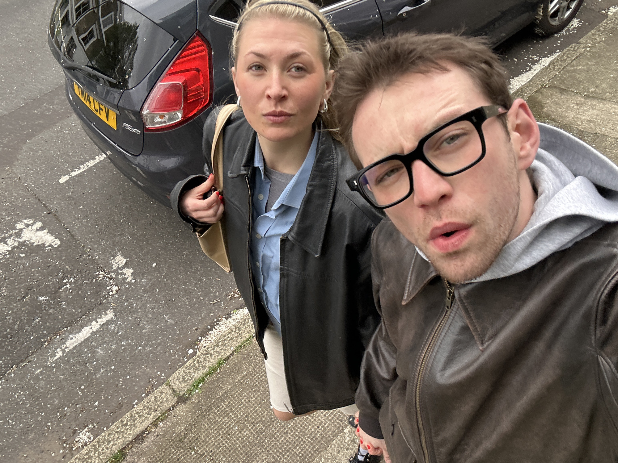 A man and a woman stand on a sidewalk near a parked dark grey Ford Fiesta, taking a selfie. The woman, fair-skinned with blonde hair pulled back with a headband, wears a black leather jacket over a light blue shirt and off-white pants. She carries a beige bag over her shoulder. The man, also fair-skinned, wears large black glasses, a grey hooded sweatshirt, and a brown leather jacket. The car has a yellow license plate that reads "TM14 CFV". The sidewalk is grey with white markings and scattered debris, next to a curb with green grass. The location is likely in the city of London, United Kingdom, based on the license plate.