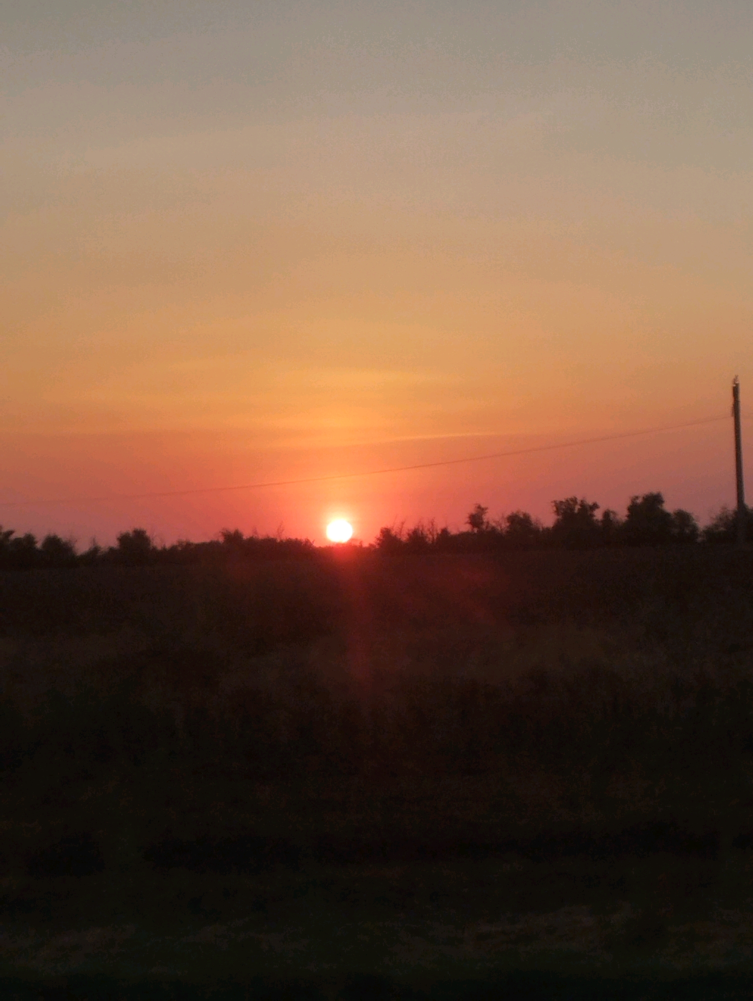 The image shows a sunset over a rural landscape. The sun is low on the horizon, casting a warm, orange glow across the sky. In the foreground, a dark silhouette of trees and a field can be seen. A utility pole stands on the right side of the image, with a wire extending across the sky. The overall scene is peaceful and serene, evoking a sense of natural beauty. The location in Kegul'ta, Russia is not discernible from the image itself, as it is a generic rural landscape.