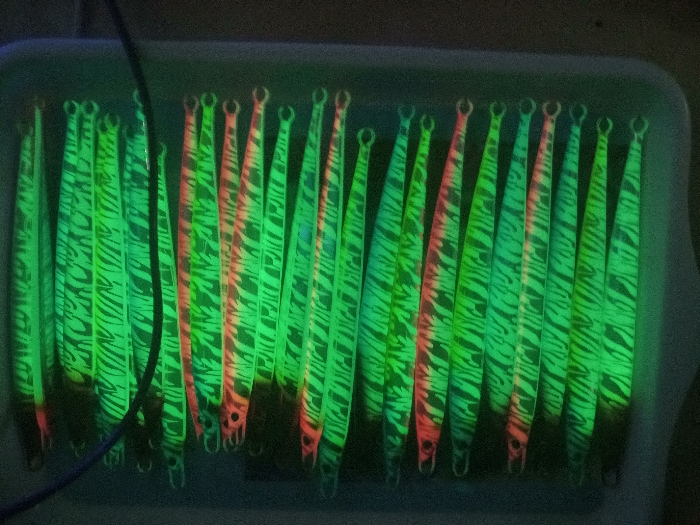 The image shows a tray of 20 fishing lures. The lures are all long and slender, with a green, white, and pink zebra pattern. They are arranged in a neat row, with their heads facing the same direction. The lures are laid in a white plastic tray with raised sides. The tray is lit from above by a fluorescent light. The background is a dark, blurry space. The image was taken in Kebumen, Indonesia, at latitude -7.662, longitude 109.647.