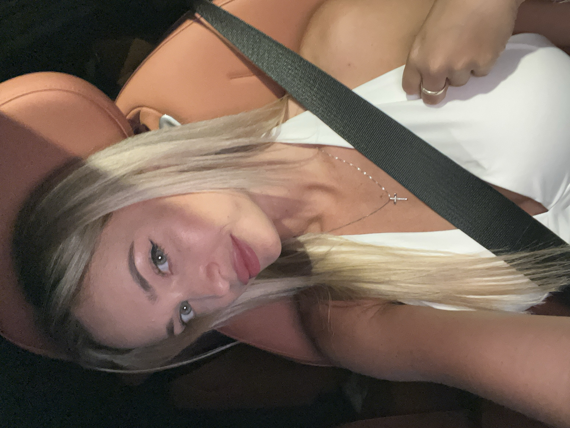 A woman with blonde hair, wearing a white top and a silver cross necklace, is sitting in a car. She is wearing a ring on her left ring finger. Her hair is long and straight, and she has light-colored eyes. The car's interior appears to be tan leather. A seatbelt is visible across her chest. The background is dark. The image is a selfie taken inside a vehicle, possibly a private car or taxi, in Sharm El Sheikh, Egypt.