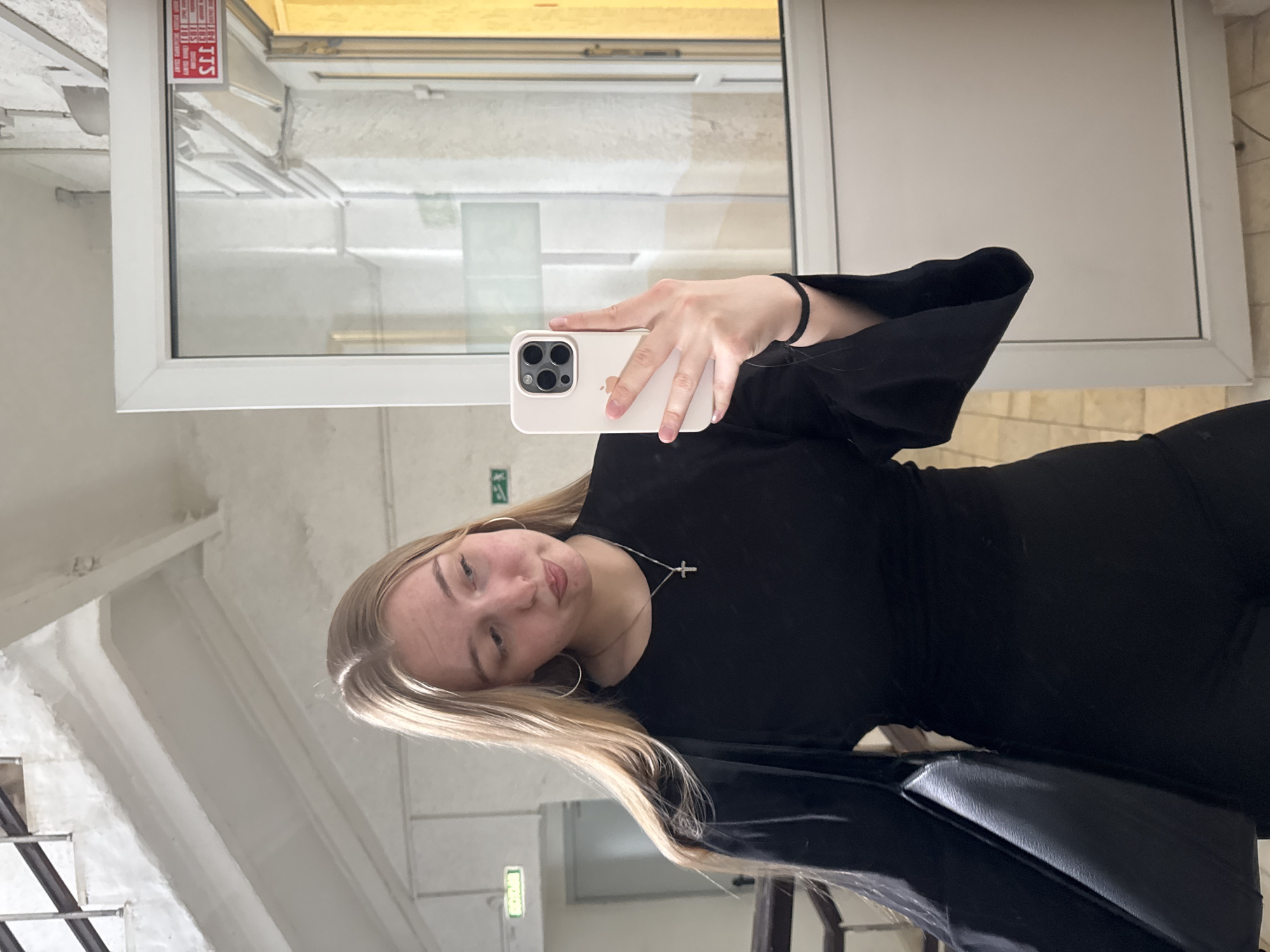 A fair-skinned, blonde woman is posing for a selfie in what appears to be a public building. She is wearing a black dress with flared sleeves, a cross necklace, and hoop earrings. She is holding an iPhone with a white case and three camera lenses. She appears to be in a stairwell with white walls and a window with white trim. Based on the sticker in the window and the architectural style, the location is likely Perm, Russia.