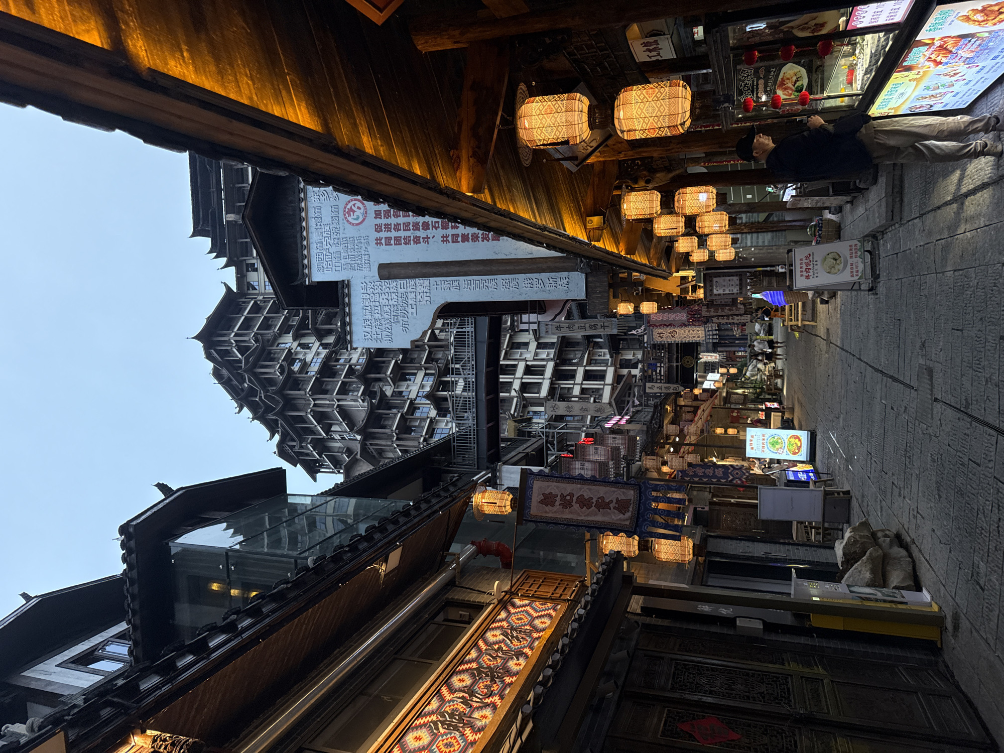 The image depicts a narrow street in Zhangjiajie, China, likely a tourist or commercial area. The street is lined with buildings exhibiting traditional Chinese architectural elements, including overhanging roofs and intricate wood carvings. Many of the buildings appear to be businesses, with illuminated signs and lanterns. Several people can be seen walking along the street. Signs display Chinese characters, and some advertisements include food-related images. The photograph is taken from a low angle, looking upwards.