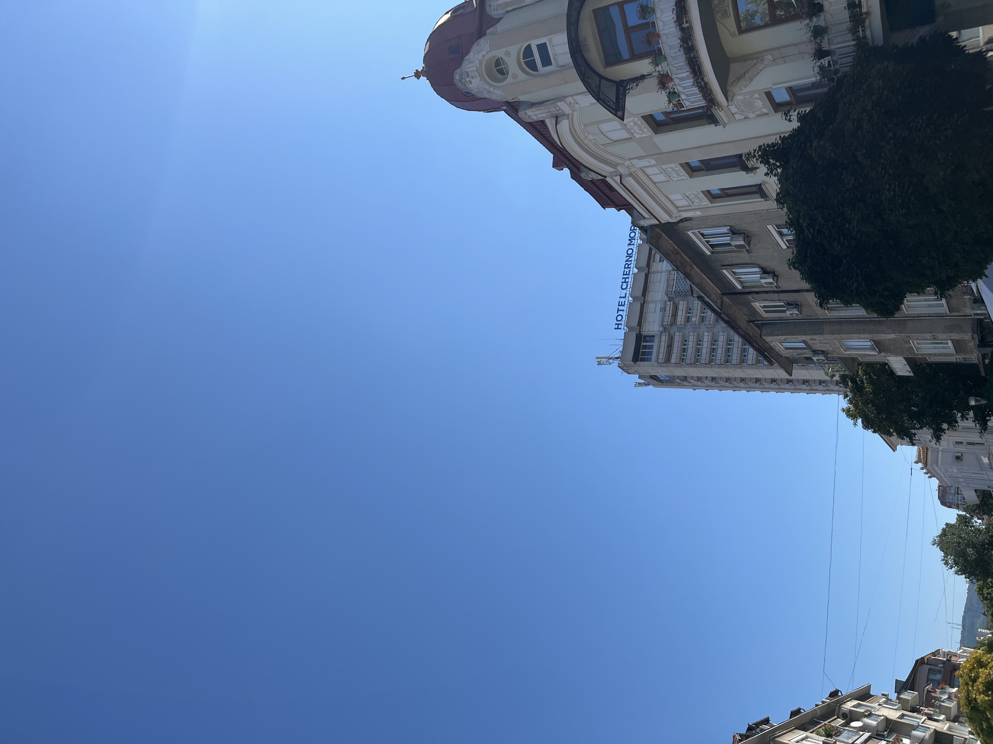 This is an outdoor, eye-level shot of an urban street scene in Varna, Bulgaria, dominated by a clear blue sky and several buildings. On the right is a multi-story building with an ornate architectural style, featuring a red domed roof, decorative facade details, and balconies with potted plants. Adjacent to it, to the left, is a modern high-rise building with the words "HOTEL CHERNO MORE" illuminated in blue on its upper section. Further to the left, other residential buildings with balconies and air conditioning units are visible. Trees are interspersed between the buildings, and power lines stretch across the frame. The composition emphasizes the contrast between the older, decorative architecture and the more contemporary structures, set against a bright, cloudless sky.