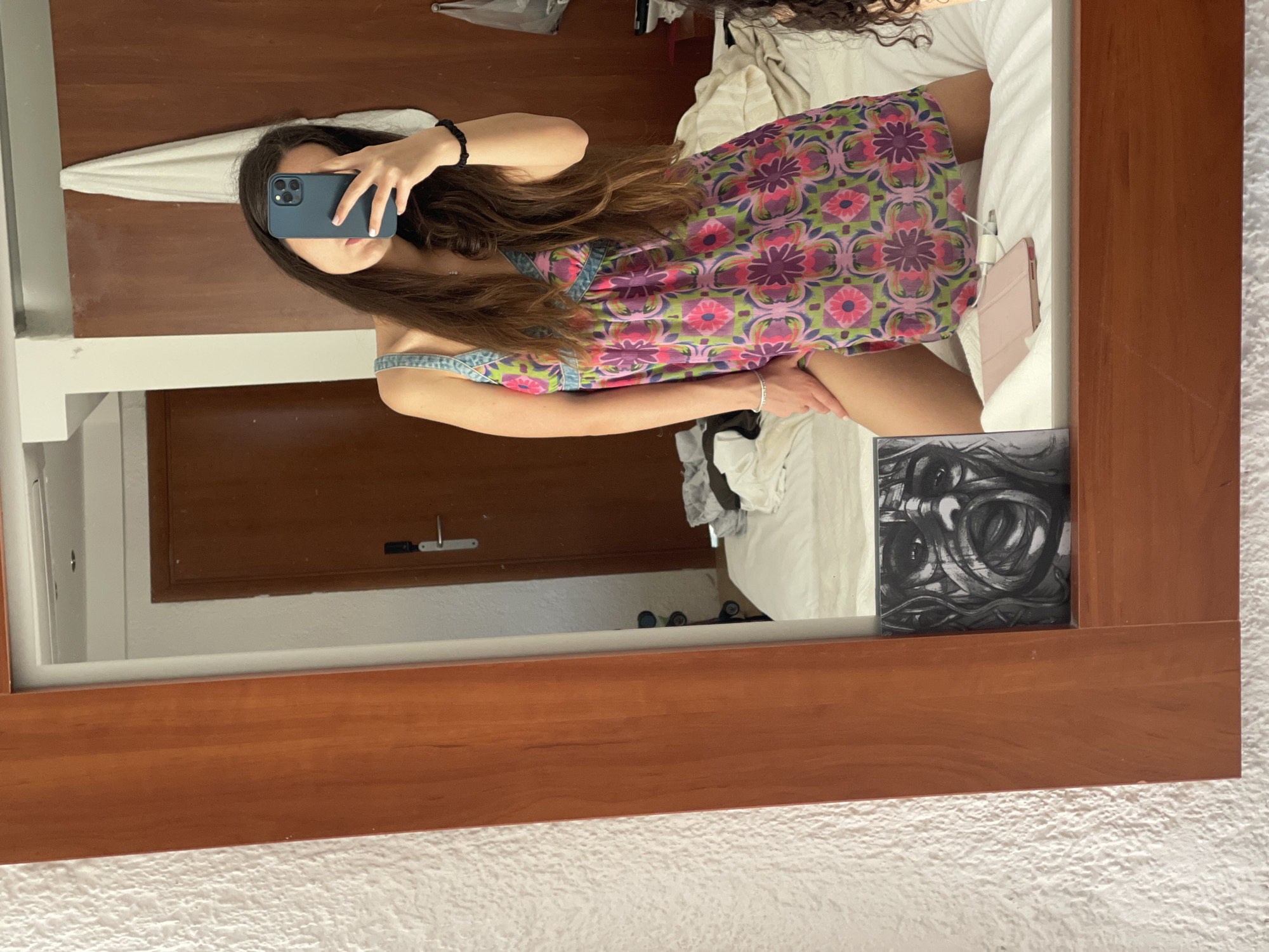 A mirror selfie captured indoors in Nea Makri Municipal Unit, Greece, shows a woman in a floral dress lying on a bed. She holds a blue phone in her left hand, partially obscuring her face. The background reveals a wooden door, a white towel, and part of a bed with white bedding. An abstract black and white artwork rests on the bedside table. The mirror's frame is made of wood, and the surrounding wall has a textured white surface. Clothes are also visible on the bed.
