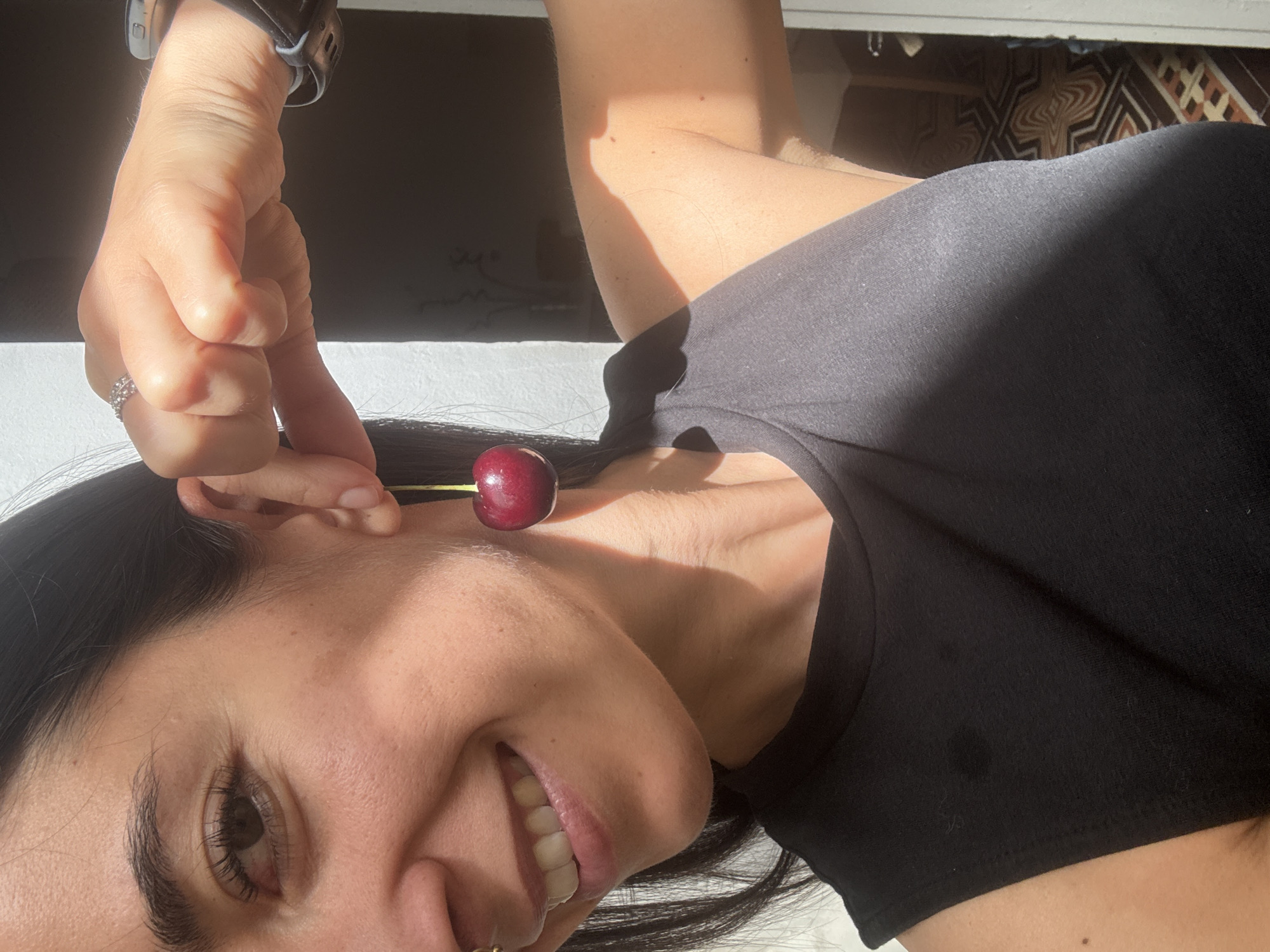 A close-up shot captures a woman's face and shoulder as she holds a cherry by its stem up to her ear. The woman is smiling, and the cherry is positioned as if it were an earring. Sunlight casts a shadow across her face and chest. The background appears to be an indoor setting, possibly a room with white walls and a patterned floor.