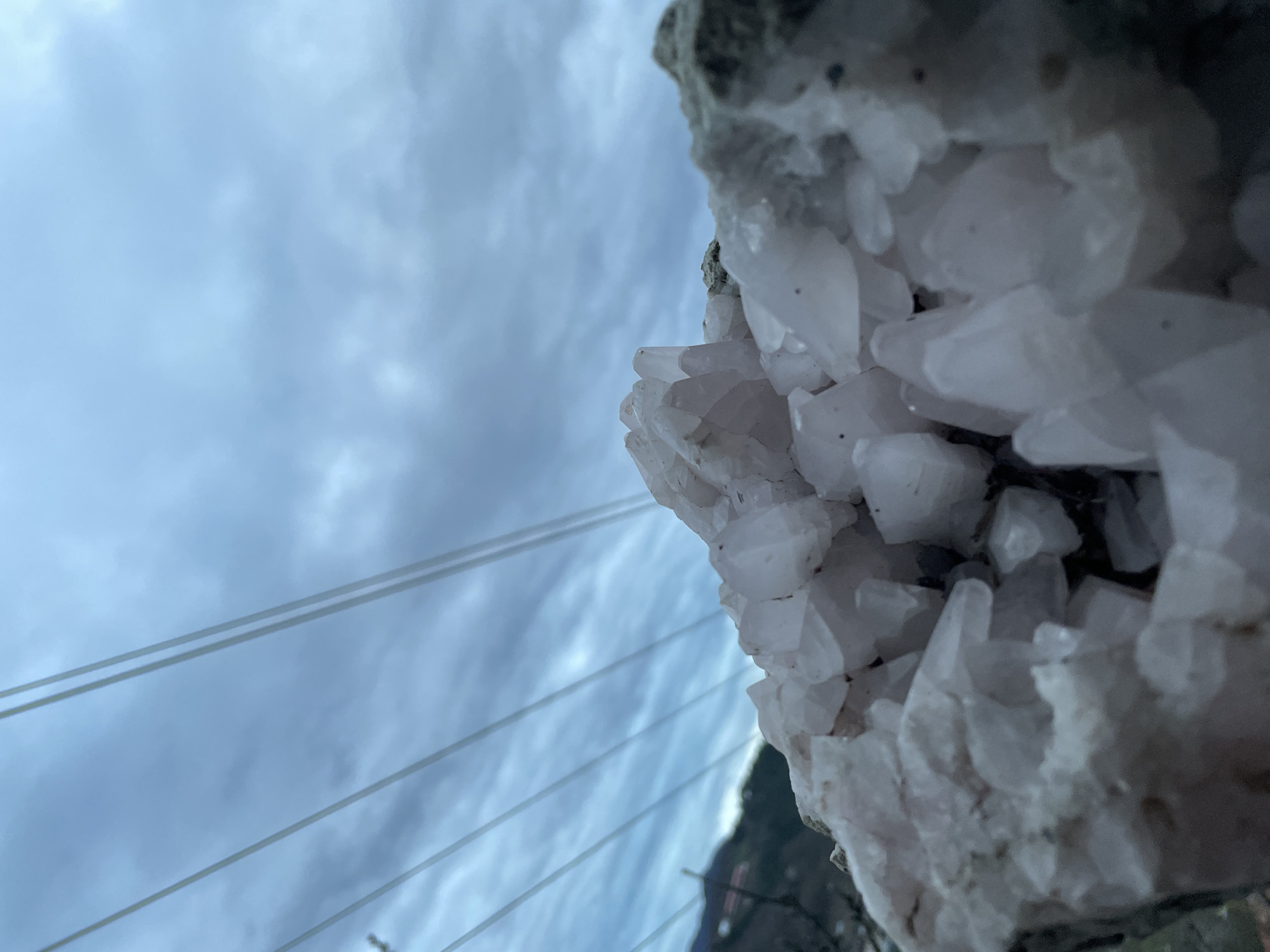 The image depicts a close-up of a quartz crystal formation against a cloudy sky. In the background, power lines are visible. The location appears to be Zlatograd, Bulgaria, indicated by the surrounding landscape elements in the image.