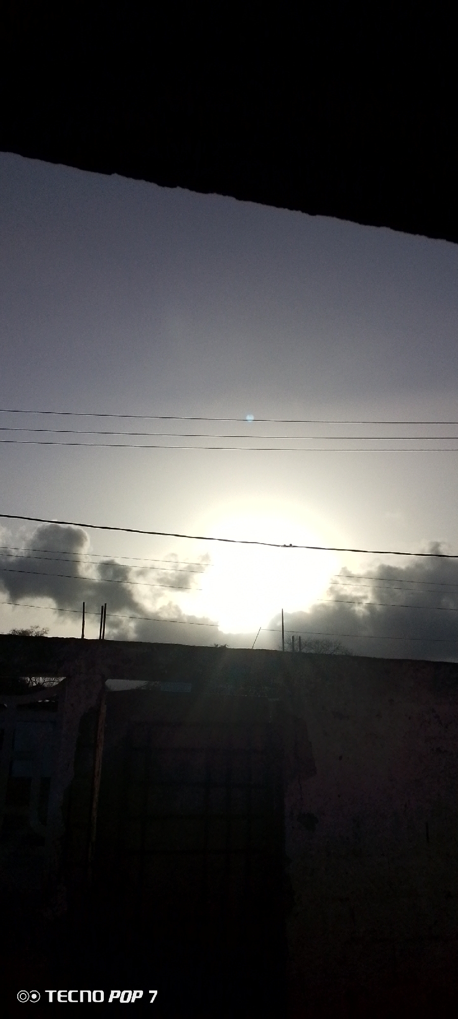 The image depicts a view from Altagracia, Venezuela, under an overcast sky with the sun partially obscured by clouds. Several power lines stretch horizontally across the upper part of the frame. A dilapidated building, possibly a house or part of a structure, stands in the foreground and is silhouetted against the bright sky. The scene is somewhat dark and lacks specific details due to the low light conditions. The camera's brand, "TECNO POP 7," is displayed in the lower left corner.