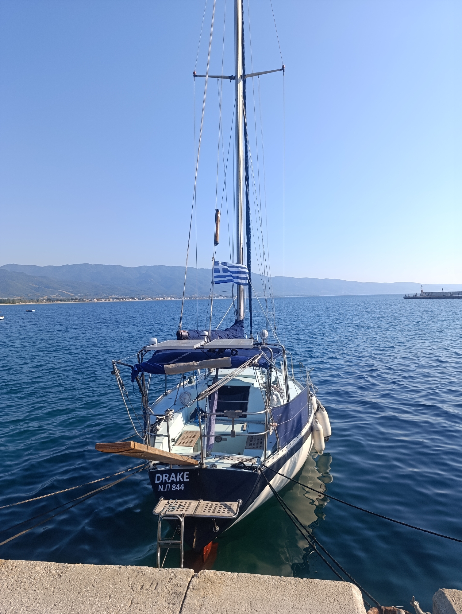 A sailboat named "DRAKE N.Π 844" is moored to a concrete dock in the sea at Stavros, Greece. The sailboat, with a blue canopy and the Greek flag flying from its mast, is seen from the stern. The water is calm and blue, reflecting the clear sky. In the background, a coastline with mountains and buildings is visible. The scene is illuminated by bright sunlight, suggesting a warm day. There are no people visible on the boat or in the immediate vicinity.