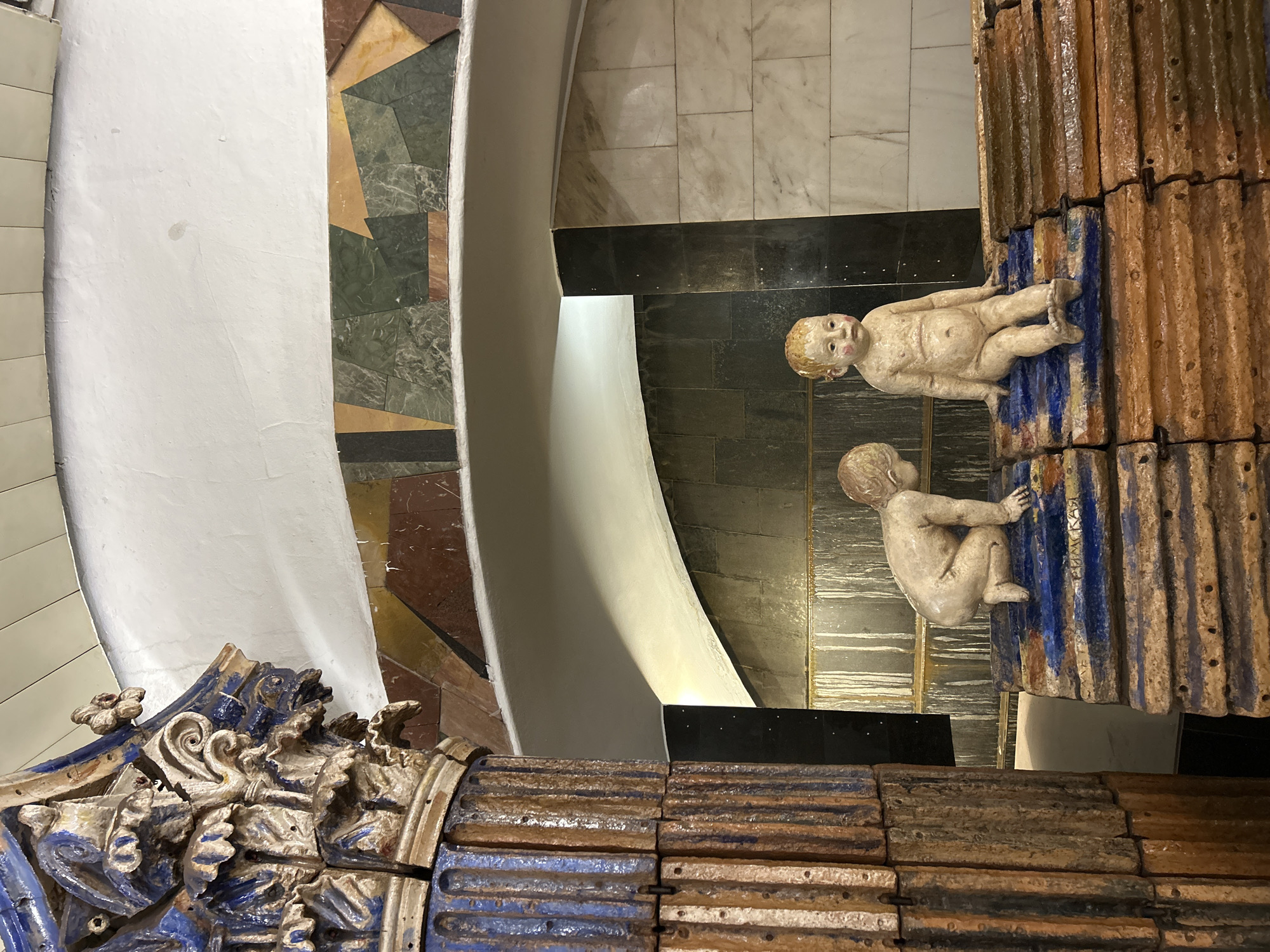 A high-angle interior shot depicts part of a building's structure featuring ornate designs and sculptures of infants. A portion of a wall with a cream-colored, textured surface is on the left side of the frame. To its right, there is a decorative strip with geometric patterns composed of various colored stones (brown, green, yellow, and grey) that are arranged in different shapes. Adjacent to the stone strip is a curved, white wall.
On the right side of the frame, there is a thick vertical structure made of stacked, grooved tiles in shades of brown and blue. Sculptures of two nude infants are affixed to this structure, appearing to climb it.
The flooring visible in the background is made of large, square tiles in light and dark grey. A black, rectangular panel is positioned between the tiled floor and the white wall.
In the bottom-left corner, there is an ornate, blue, and white sculptural element. The overall lighting appears to be soft and diffused.
The location is Unknown, Unknown.