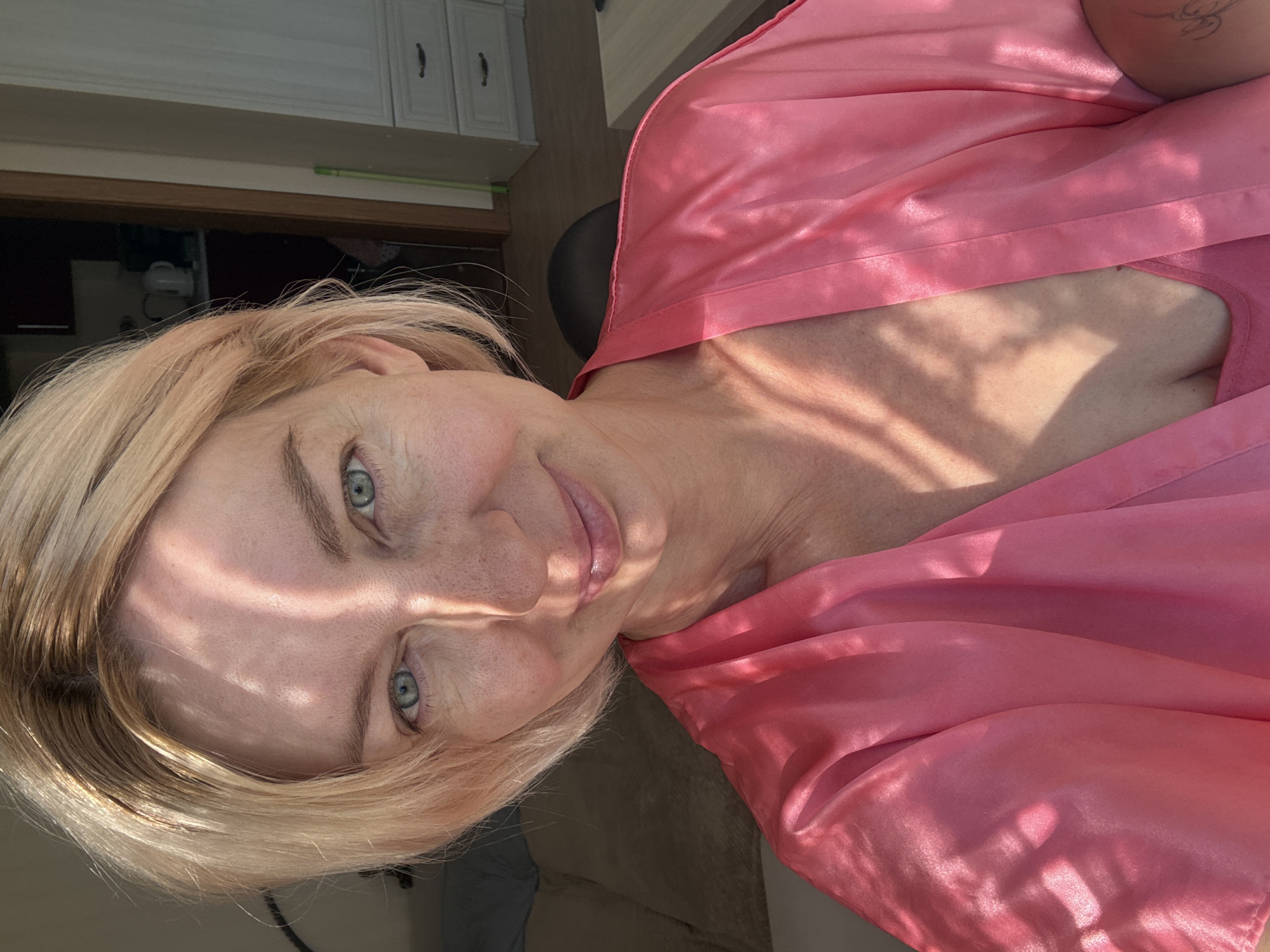 An over-the-shoulder, close-up selfie of a fair-skinned woman with short blonde hair and blue eyes. She is wearing a pink silk robe over a pink camisole. The woman is looking directly at the camera with a slight smile. Sunlight is casting dappled shadows across her face and upper body. The background is blurred, but suggests an indoor environment with furniture and a doorway to another room. The image does not provide enough information to determine the location in Sochi, Russia.