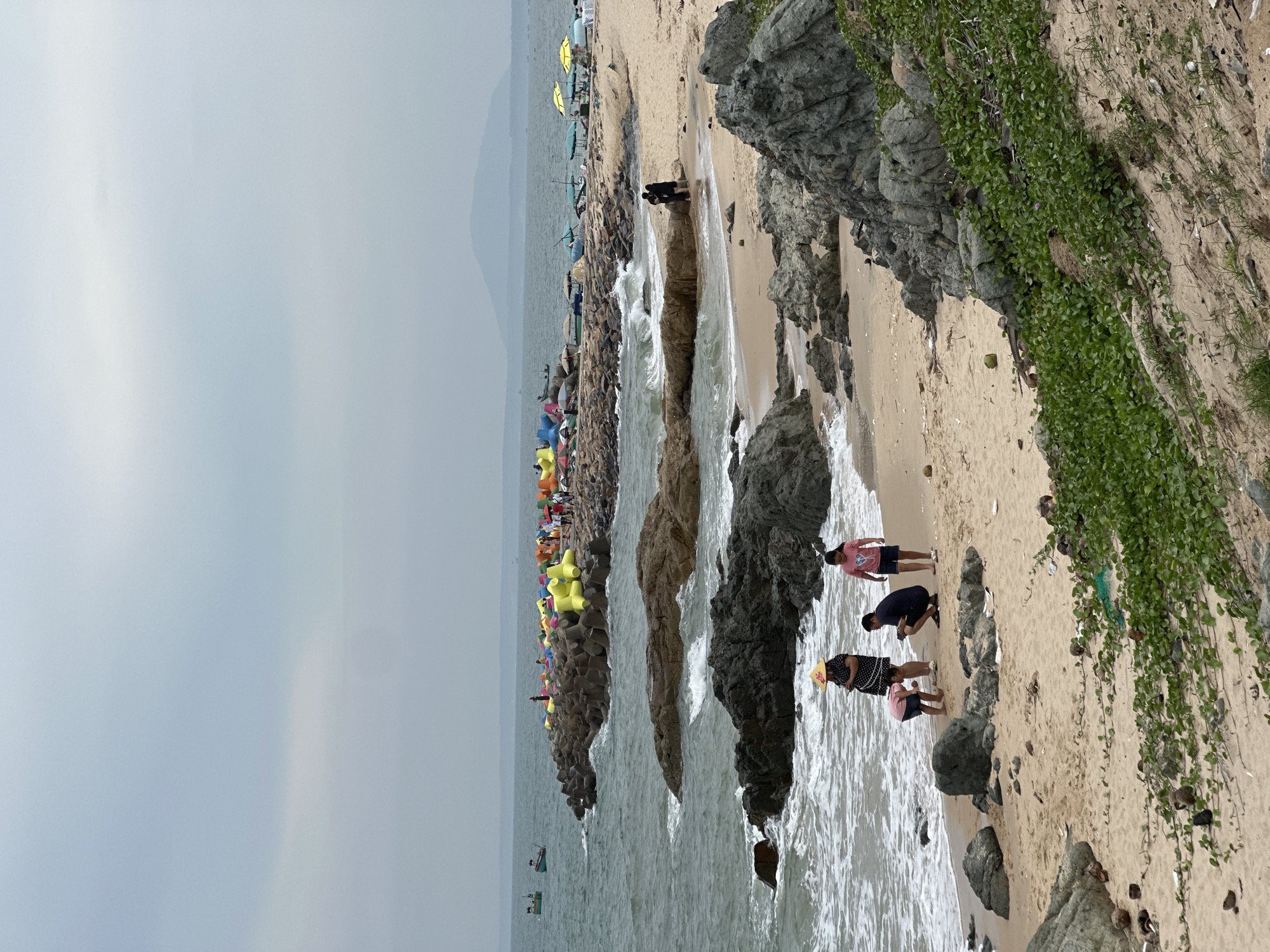 The image depicts a beach scene in Phu Thuy Ward, Vietnam, characterized by a sandy shore, rocky outcrops, and a calm sea under an overcast sky. In the foreground, a group of four individuals, including two adults and two children, are interacting on the beach near the water's edge. One adult, wearing a wide-brimmed hat and a patterned dress, appears to be looking at her phone. A child in a pink t-shirt and shorts is crouched down, possibly examining something on the sand. Another adult, dressed in dark clothing, is kneeling and looking towards the child. A second child, in a pink t-shirt and blue shorts, stands nearby. In the middle ground, a rocky breakwater extends into the sea, adorned with brightly colored, geometric structures that appear to be inflated floats or decorative elements. Several beach umbrellas are set up on the sand behind the breakwater, suggesting a recreational area. Further out at sea, a few small fishing boats are visible. The background features a distant, hazy mountain range. The beach shows signs of natural debris, including coconuts and scattered litter, particularly in the lower right quadrant of the image where vegetation is growing on a sandy embankment.