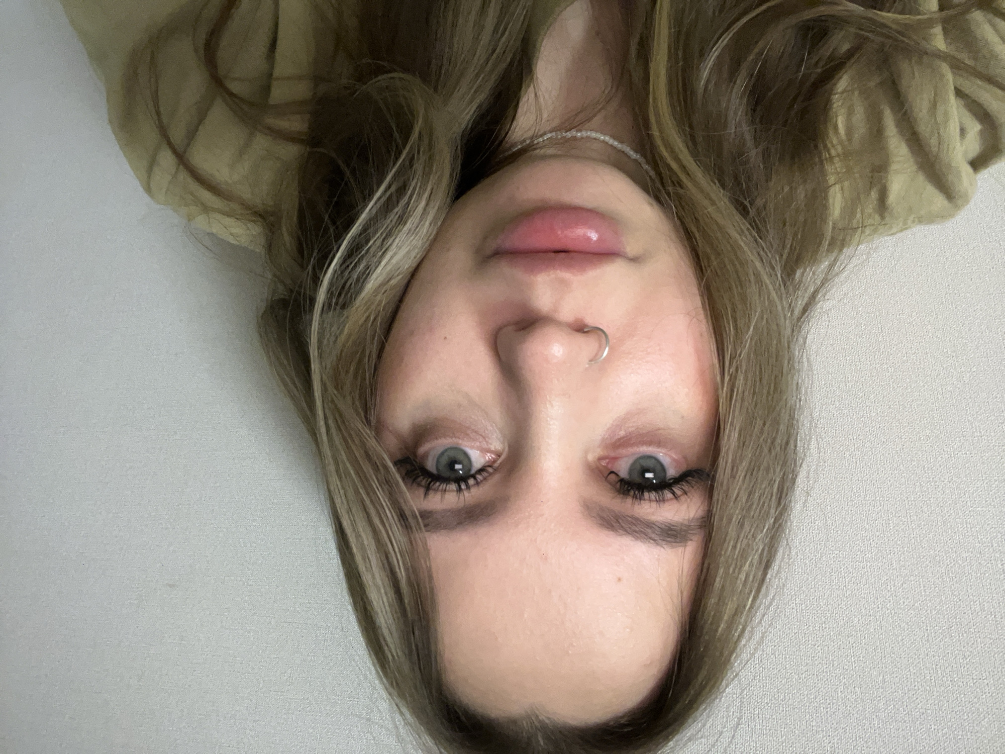 A close-up portrait shows a woman's face, upside down, with light hair. The woman has fair skin, light grey eyes, and a silver nose ring. She is wearing what appears to be a light brown top. The background is a plain light grey or white wall. The location is not discernible from the provided image.