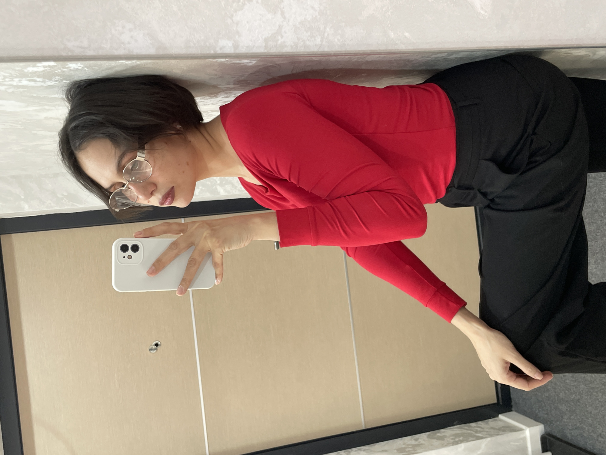 A person is taking a selfie in front of a mirrored wall. The person is wearing round eyeglasses, a long-sleeved red shirt, and black pants. The phone is white with multiple lenses. The environment appears to be indoors, possibly a bathroom or dressing area. There is no information to definitively identify the location as Izhevsk, Russia.