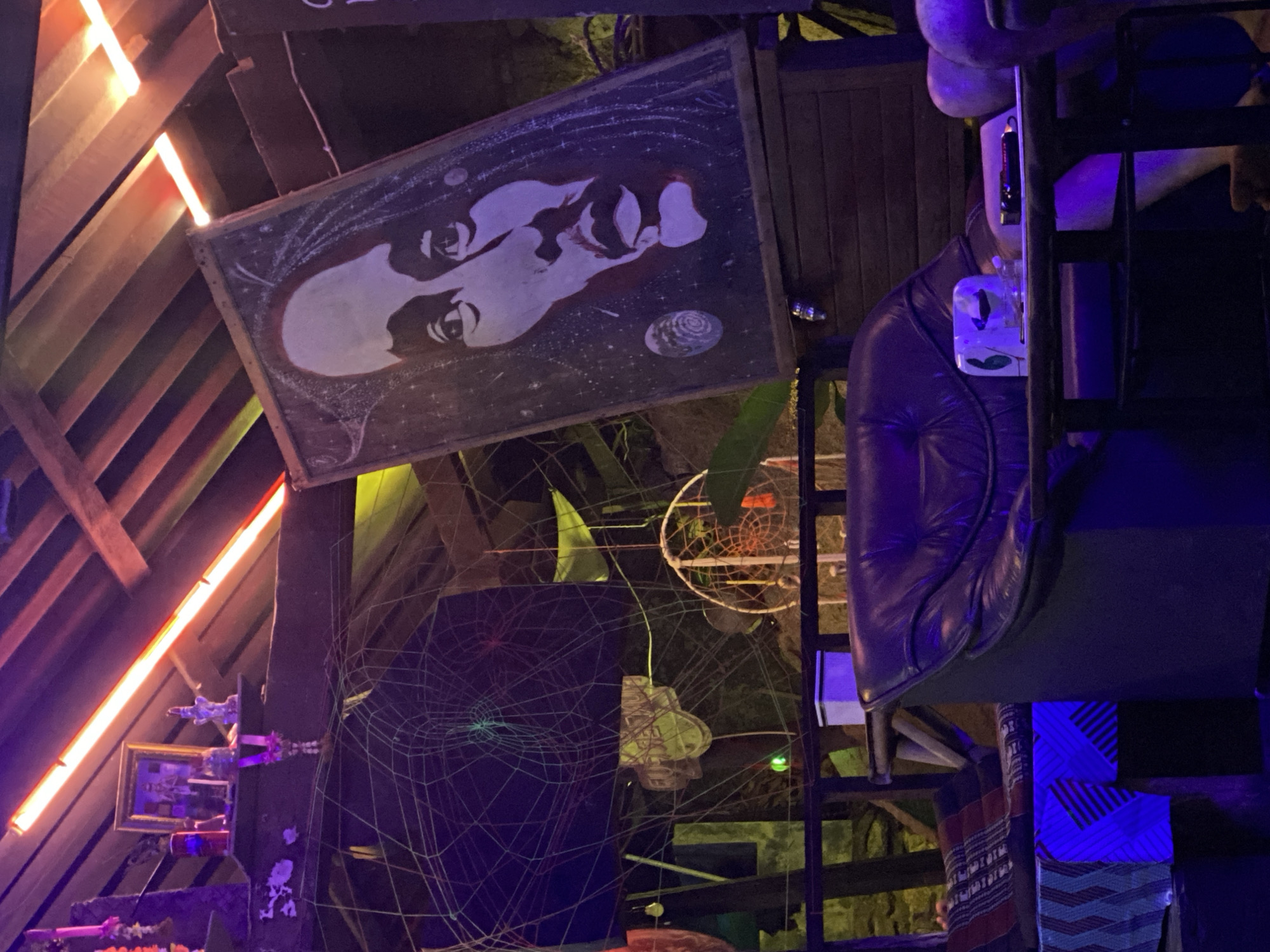 The image, taken in Baan Don Sai, Thailand, shows an eclectic interior space. At the top left, a slanted, partially visible wooden structure with orange lighting runs along its edges. A large, rectangular painting with a black background and a white, stylized face is mounted on the wall. Underneath, there's a shelf holding decorative items, including a small framed picture and several trinkets. A large, dark spiderweb-like structure occupies the lower left quadrant. To the right, a dreamcatcher hangs suspended in the background. In the foreground, a purple leather chair is positioned on a metal frame, with a small, white object attached to the chair's side. Below the chair, patterned fabrics are draped over the metal structure. The overall lighting is dim and colorful, with a mix of natural and artificial light sources.
