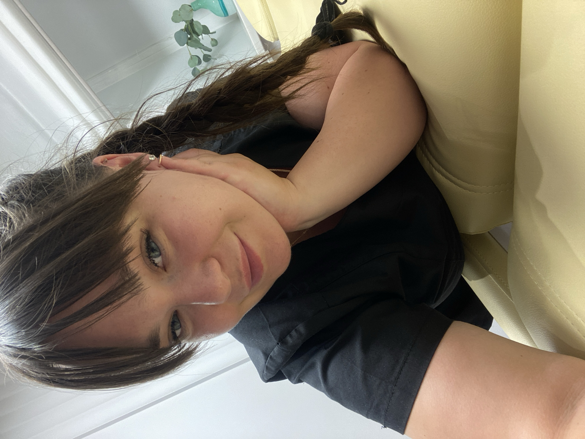 This media file is a selfie of a woman taken indoors, likely in a salon or spa setting, given the presence of a beige leather chair and a plant with eucalyptus leaves. The woman has long, dark brown hair styled in a braid and is wearing a black top. Her left hand is resting on her cheek, and she is looking directly at the camera with a relaxed expression. The lighting suggests it is daytime. There is no visible text in the image. The scene is casually intimate and suggests a moment of self-care or relaxation.