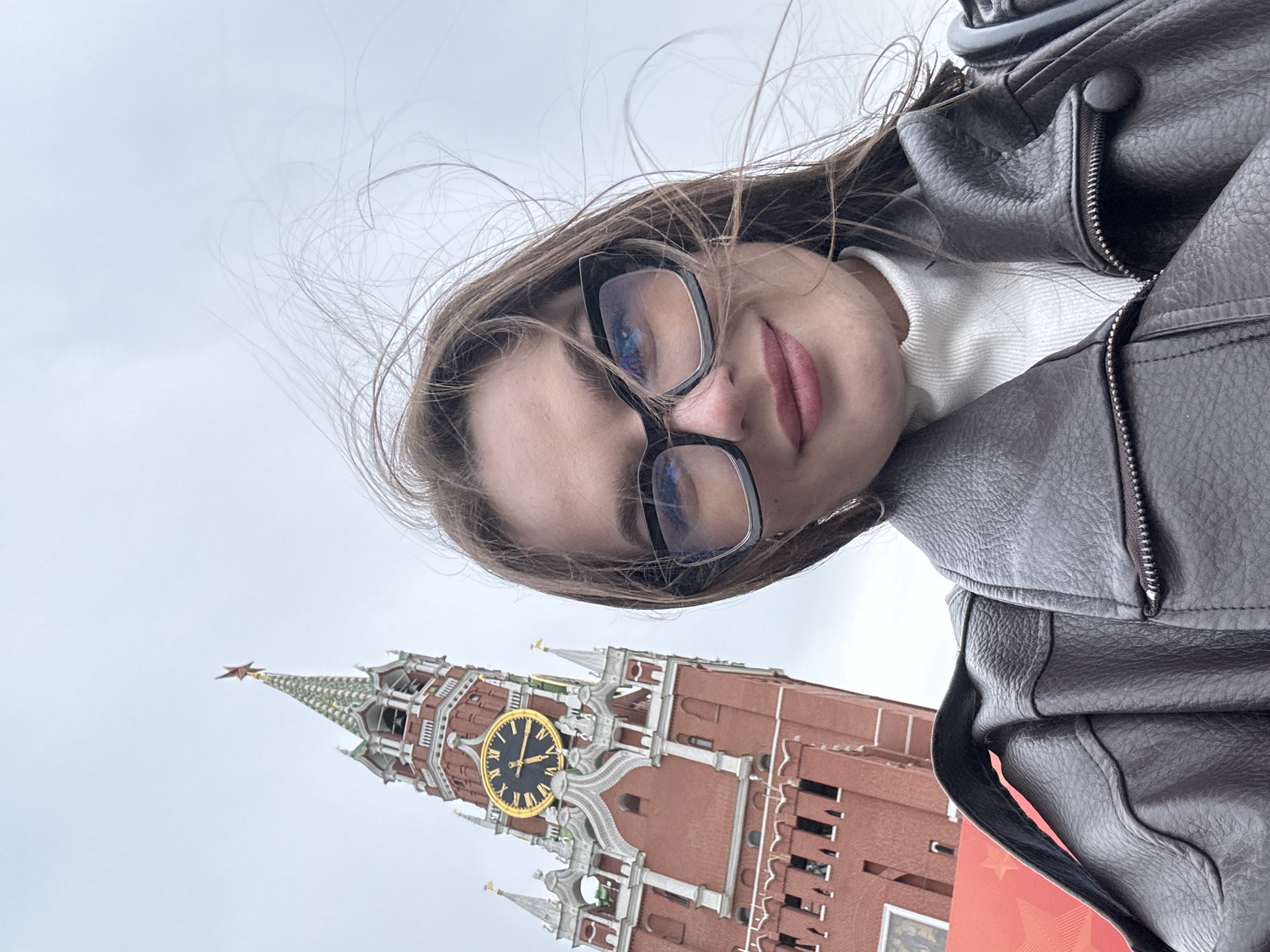 A young woman with long brown hair and glasses is taking a selfie in Moscow, Russia. In the background is the Spasskaya Tower of the Moscow Kremlin, with a gold-faced clock prominently displayed. The sky is overcast and gray. The woman is wearing a brown leather jacket and a white turtleneck sweater. Her hair is slightly windblown.