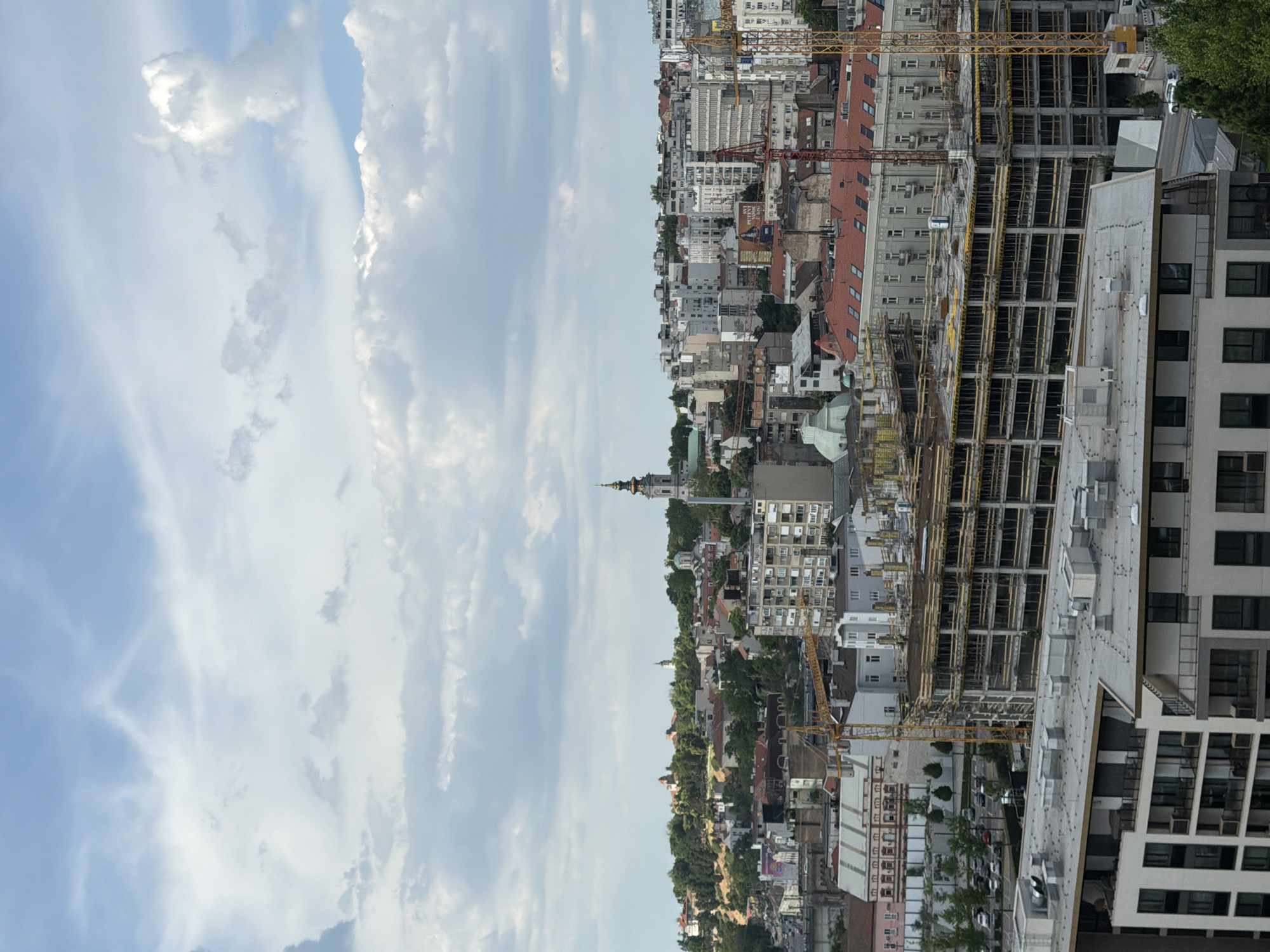 This image shows a skyline view of Belgrade, Serbia, featuring both older and newer buildings and construction sites. The sky is partially cloudy. There are construction cranes present among the buildings. In the distance, a distinct church spire is visible. The image suggests an urban environment with ongoing development.