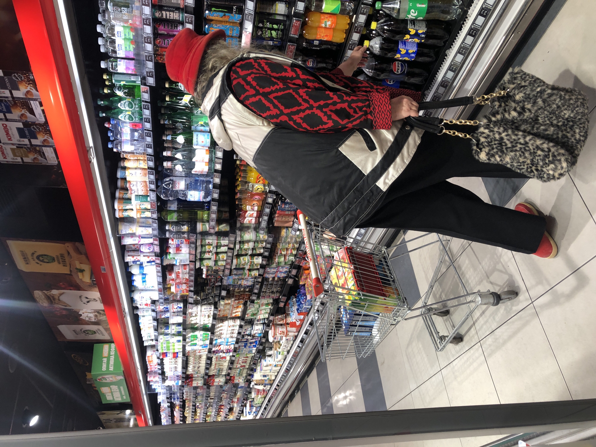 The image shows a person shopping in a supermarket aisle. The person, wearing a red hat, a black and red patterned jacket, and black pants, is reaching into a refrigerated display case filled with beverages. The person is holding a purse with a gold chain. A shopping cart containing various items is positioned next to them. The aisle is stocked with numerous products, including drinks, dairy products, and coffee, all displayed on shelves behind clear doors. The supermarket setting is brightly lit, and the floor is tiled in a light grey and white pattern. Based on the OCR text found on product labels, this is likely a store in Sofia, Bulgaria.