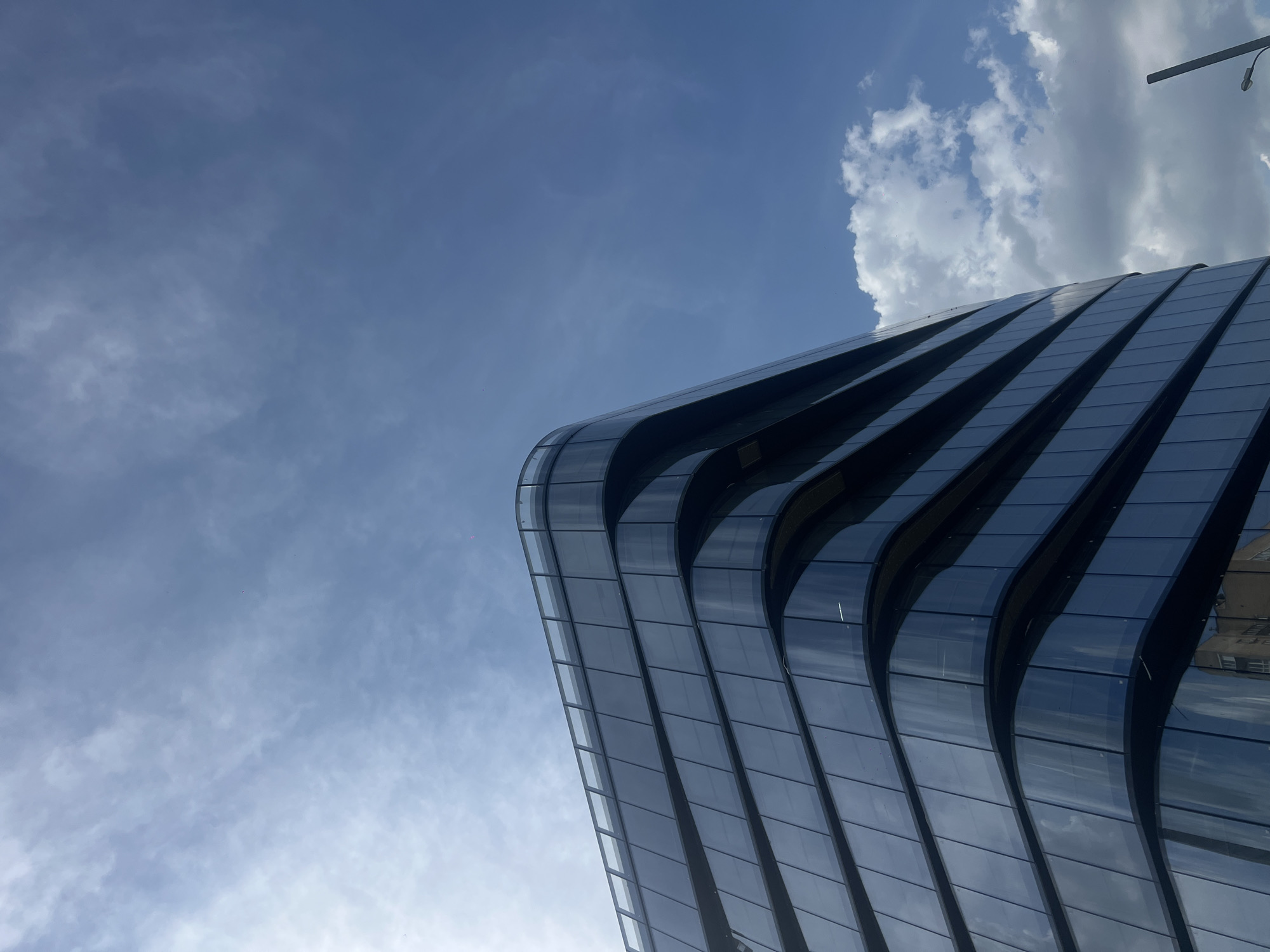 The image shows a low-angle view of a modern, curved glass building against a partly cloudy blue sky. The building's design features horizontal bands that create a wave-like effect. The glass panels reflect the sky. The sky is blue with wispy clouds. The building is located in Moscow, Russia.